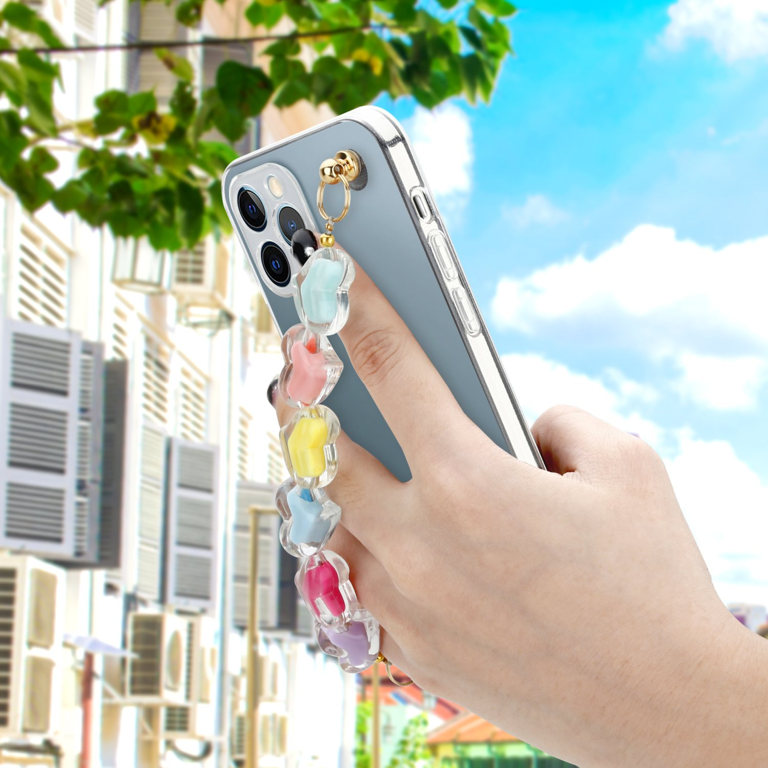 Eine Hand hält ein Telefon mit einem Band aus bunten Blumenanhängern, mit Gebäuden und Himmel im Hintergrund.
