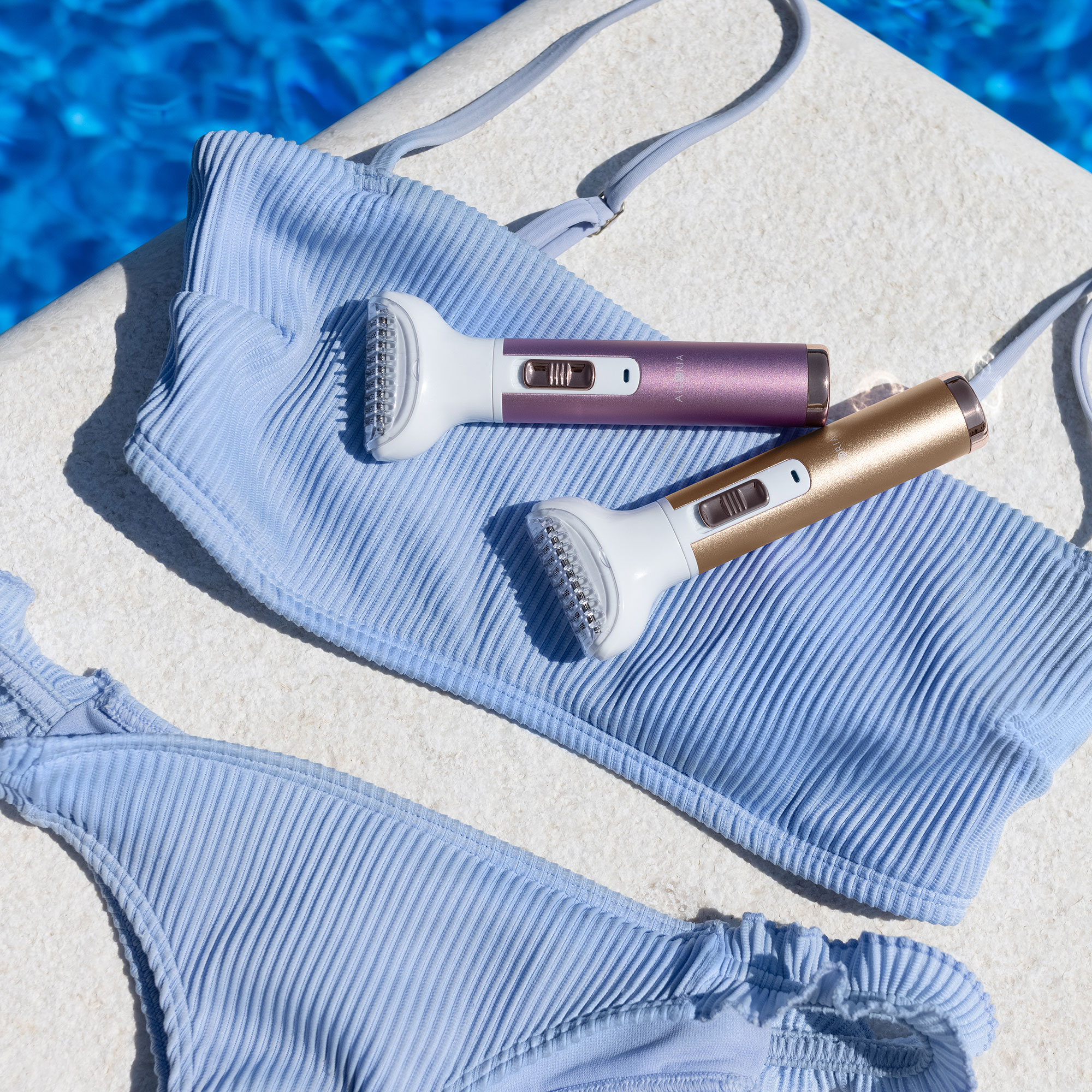 Dos depiladoras y un bikini sobre una superficie blanca junto a una piscina. Uno dorado, otro morado.