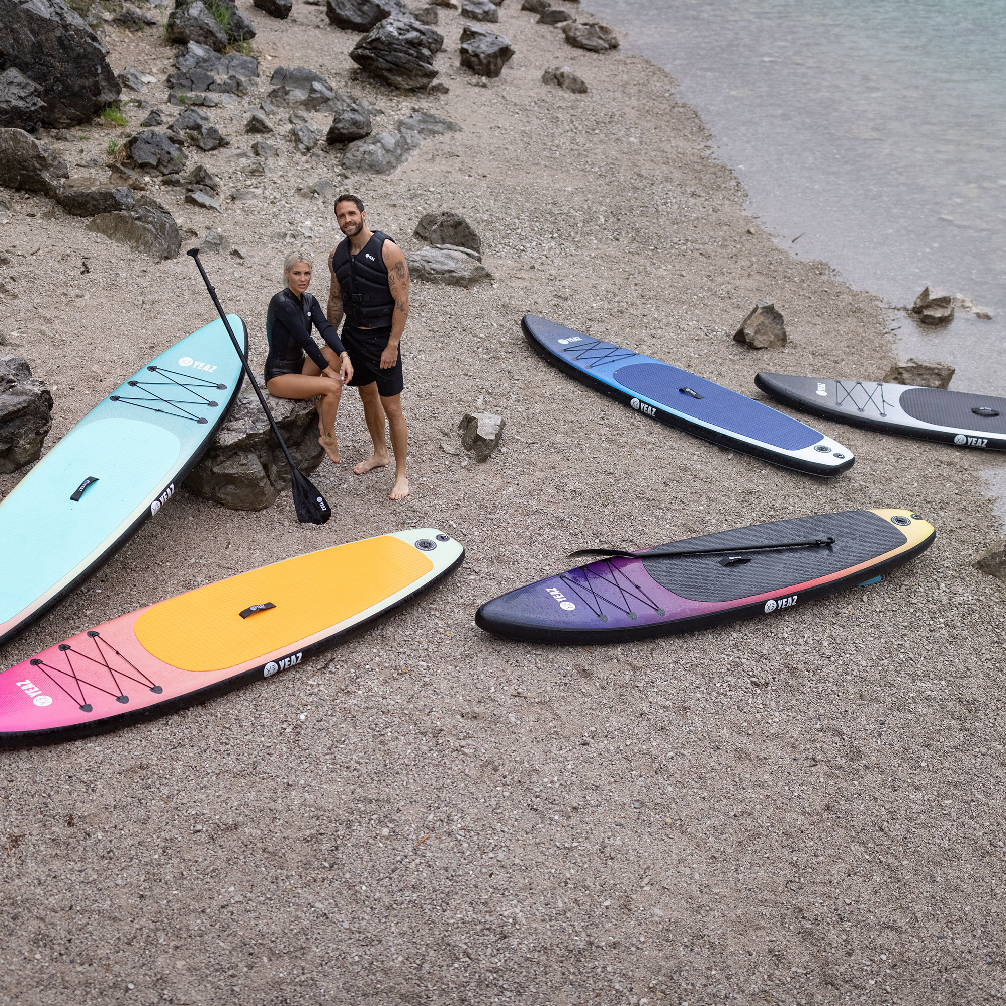 Zwei Personen stehen in der Nähe von Stand-Up-Paddleboards an einem Strand. Es gibt mehrere Boards in verschiedenen Farben.