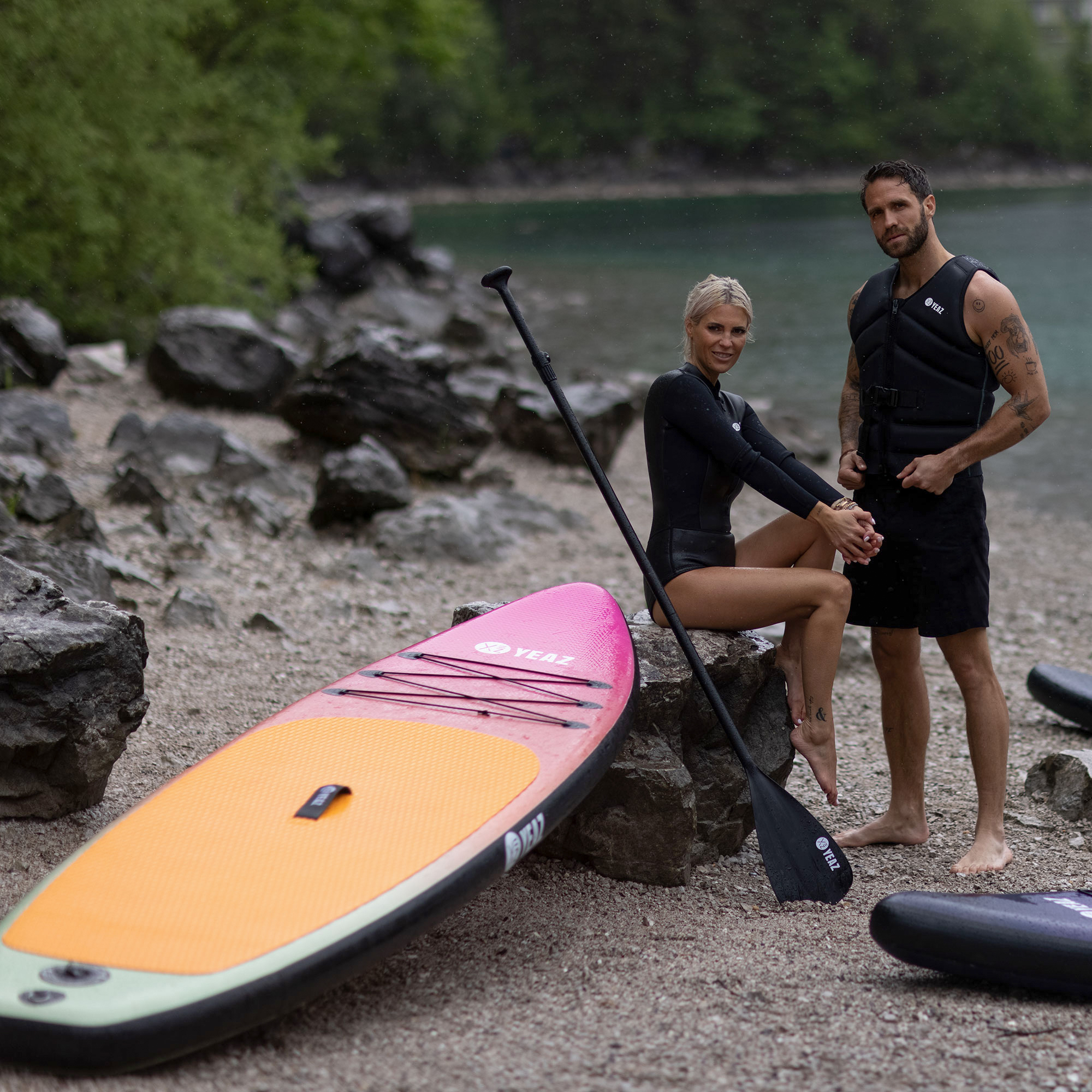 Zwei Personen am Strand mit einem Paddleboard. Mann in Weste, Frau im Neopren.