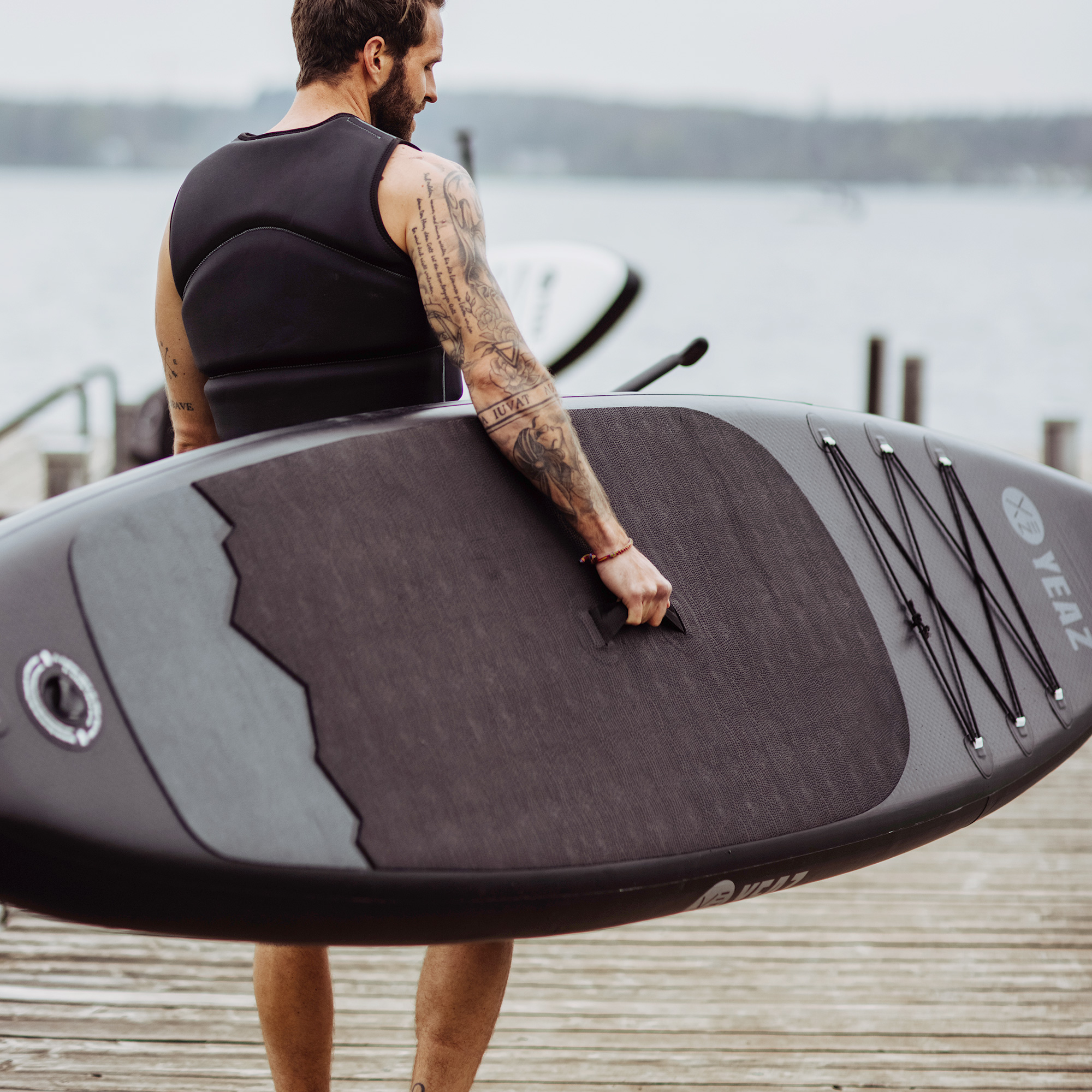 Ein Mann trägt ein Paddleboard auf einem Steg. Der Hintergrund zeigt Wasser und ein entferntes Ufer.
