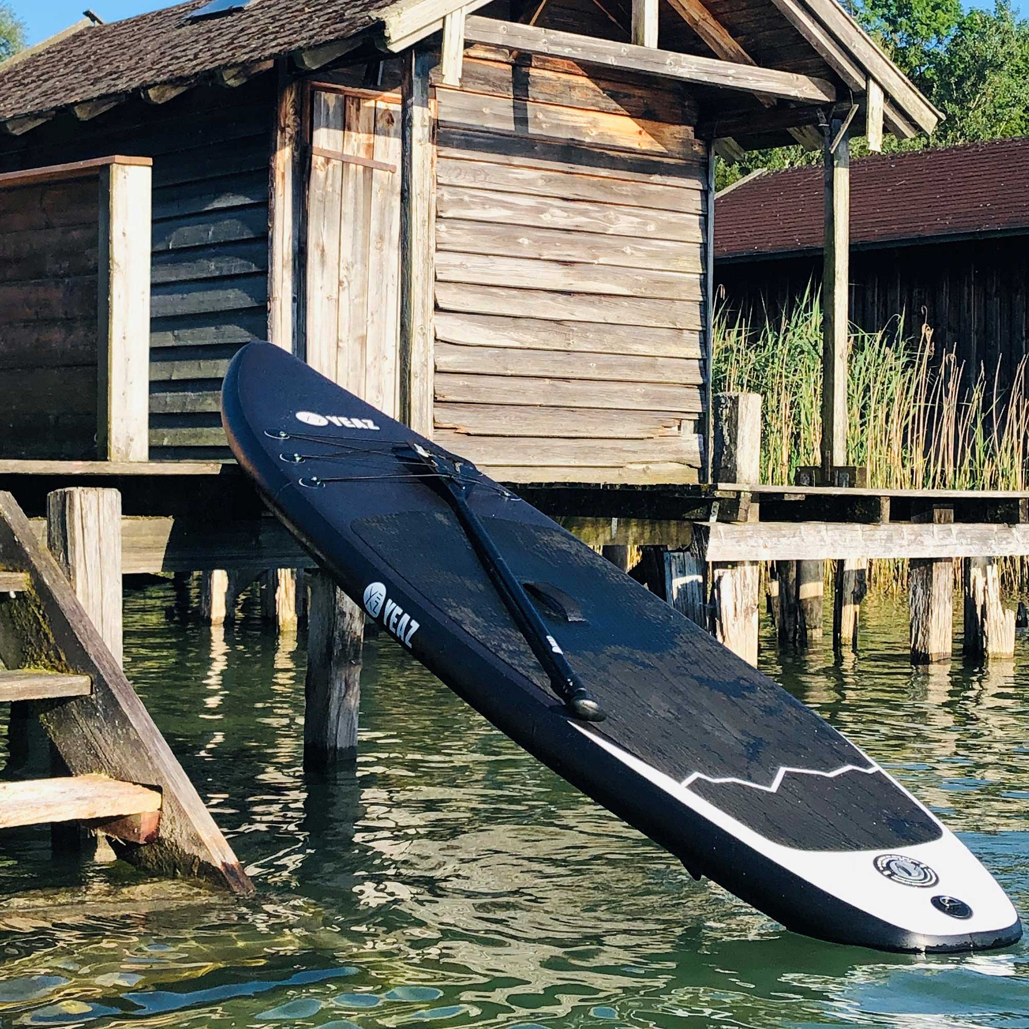 Schwarzes Paddleboard lehnt an einem Holzsteg, mit See und Holzgebäude im Hintergrund.