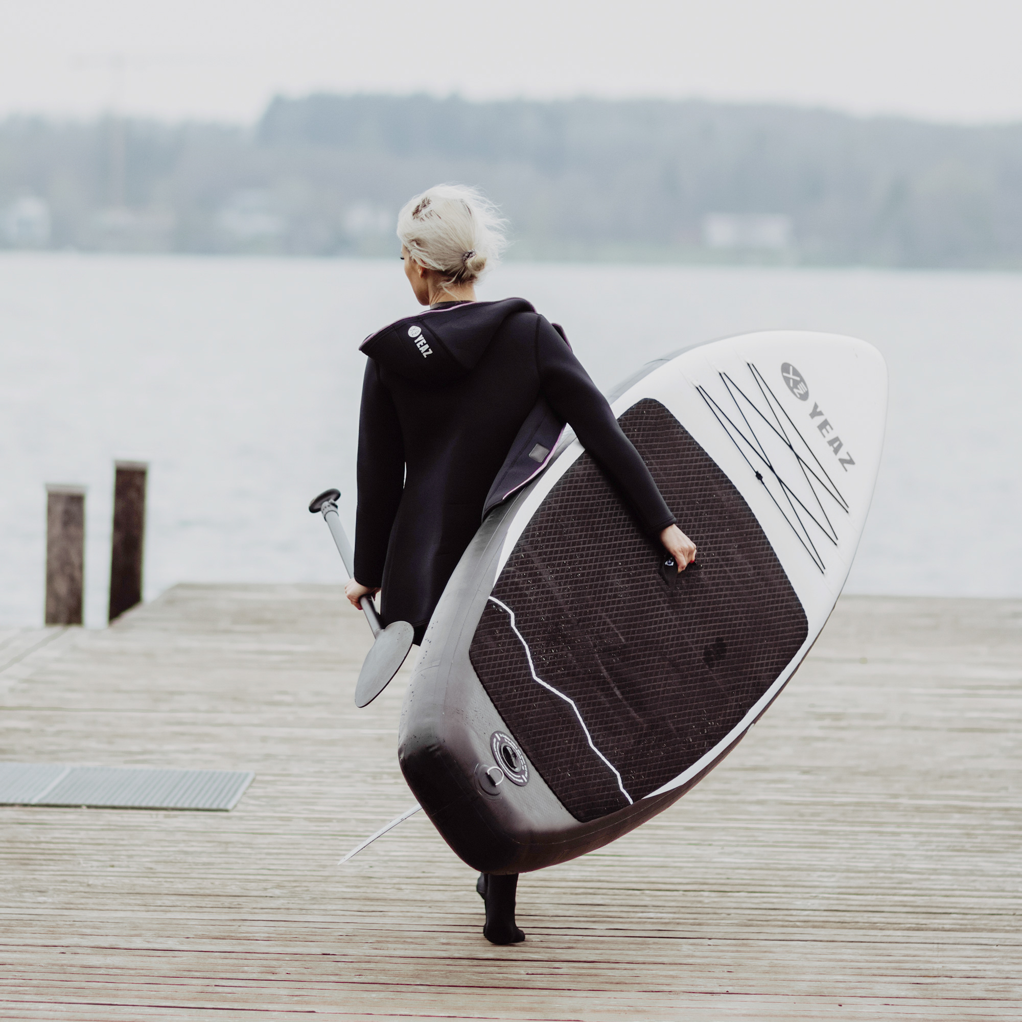 Frau in schwarzem Neoprenanzug trägt ein Paddleboard auf einem Holzsteg, See im Hintergrund, bedeckter Himmel.