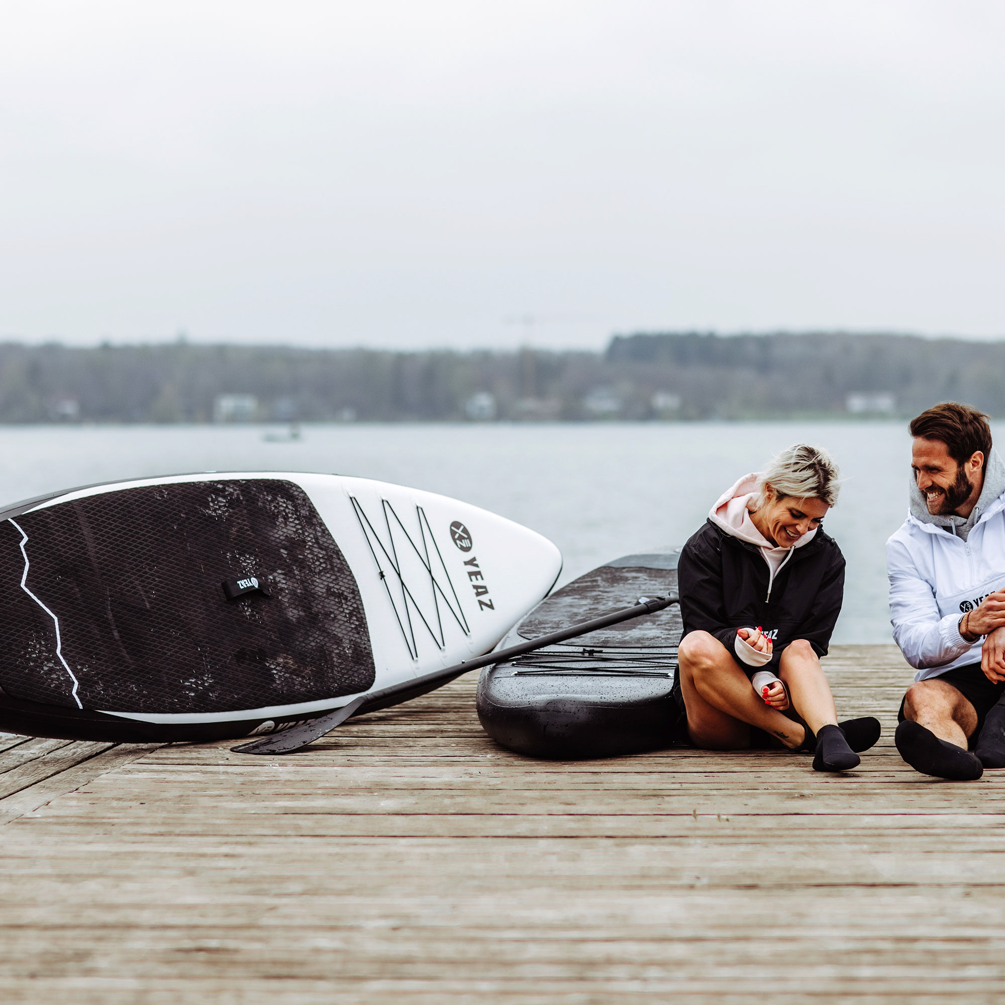Zwei Personen sitzen auf einem Holzsteg mit Paddleboards. Wasser und Bäume im Hintergrund.
