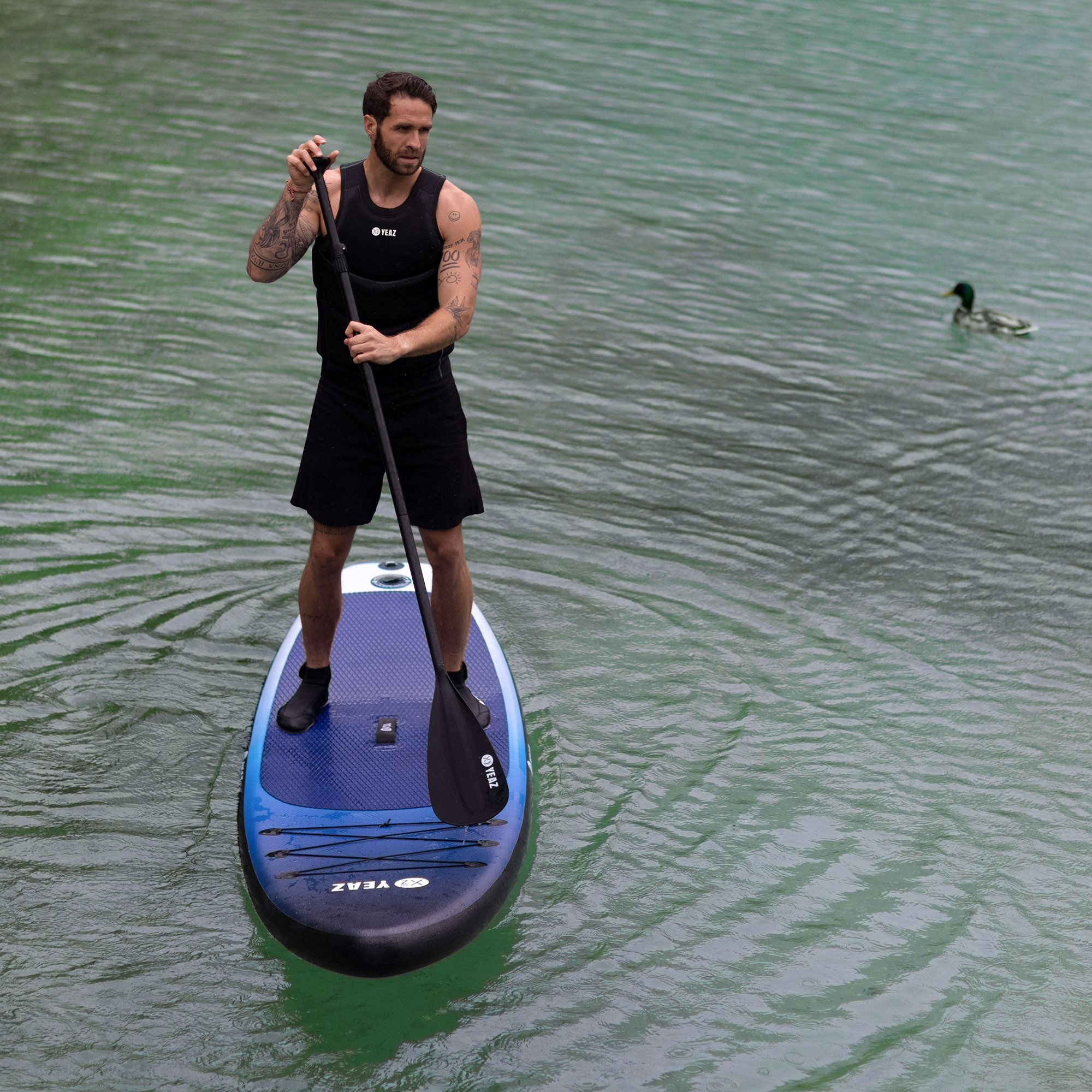Mann paddelt auf See, trägt schwarze Shorts und Weste. Ente schwimmt in der Nähe.
