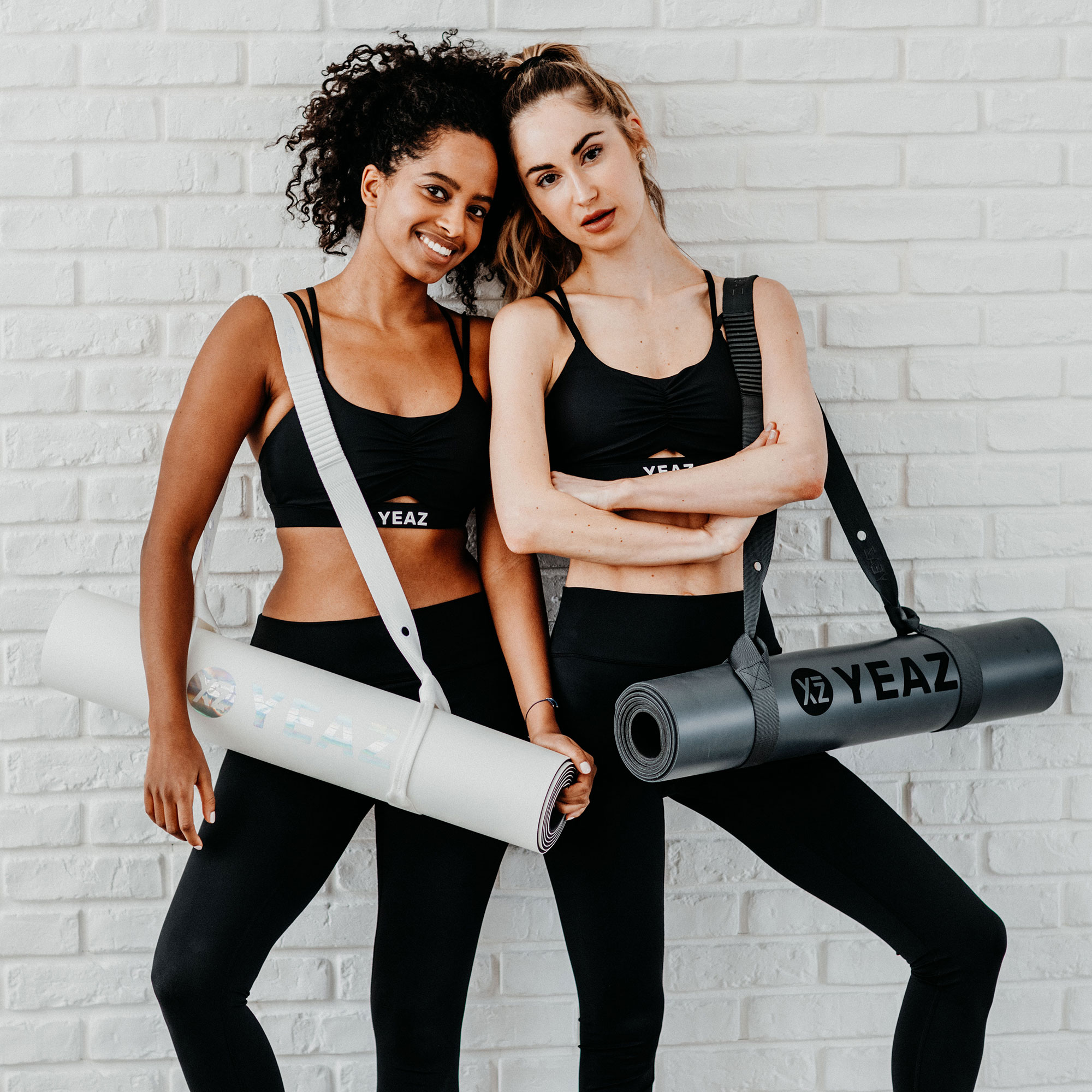 Zwei Frauen mit Yogamatten. Sie lächeln, tragen Sport-BHs und Leggings. Hintergrund Backsteinmauer.