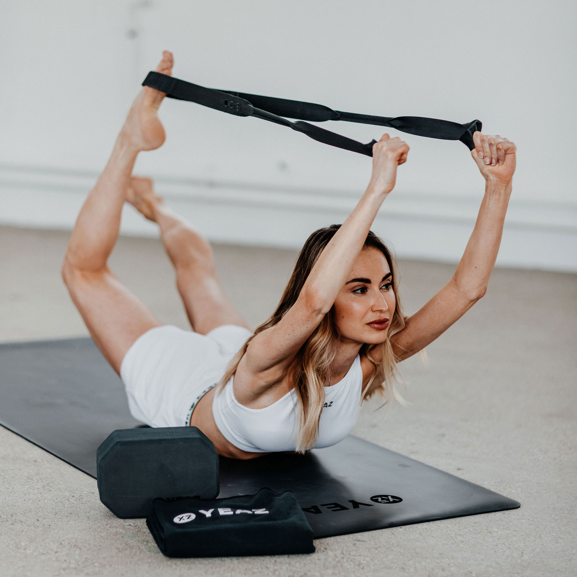 Frau in weißem Sport-BH und Shorts macht Yoga. Sie hält einen Gurt und ist auf einer Matte.