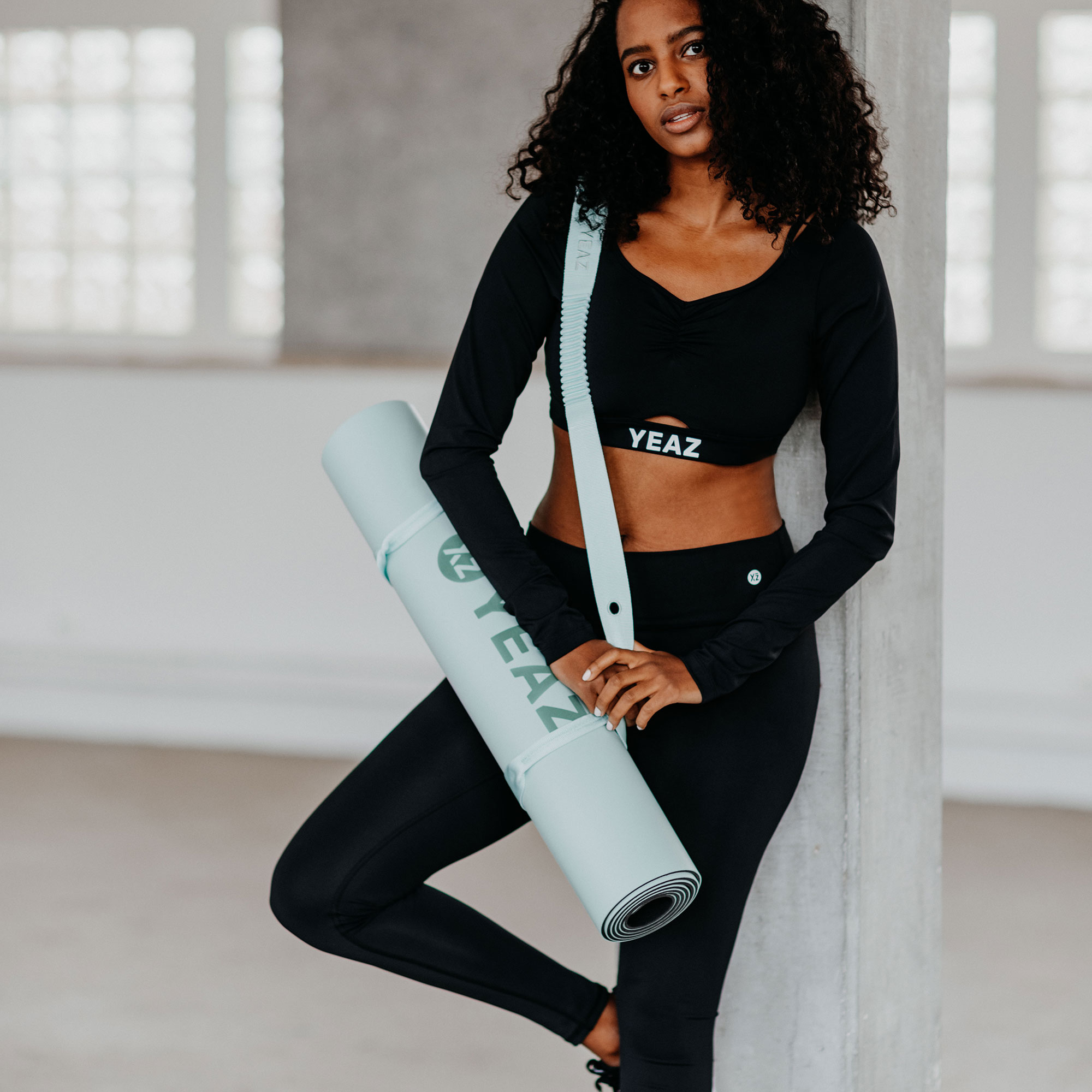 Eine Frau in Sportkleidung hält eine gerollte Yogamatte. Sie lehnt sich an eine weiße Wand.