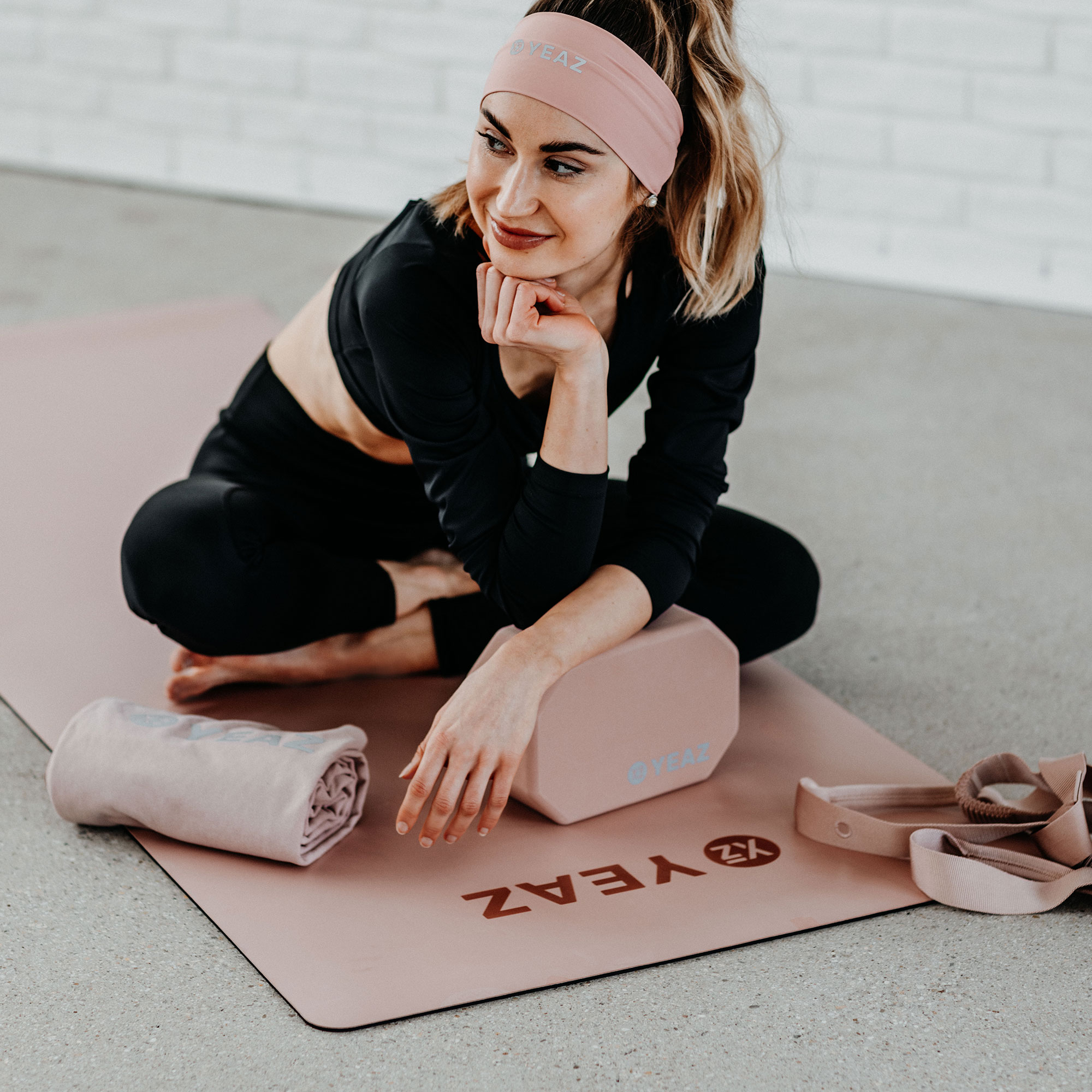 Frau mit pinkem Stirnband sitzt auf Yogamatte mit Handtuch, Block und Tasche; Marke YEAZ.