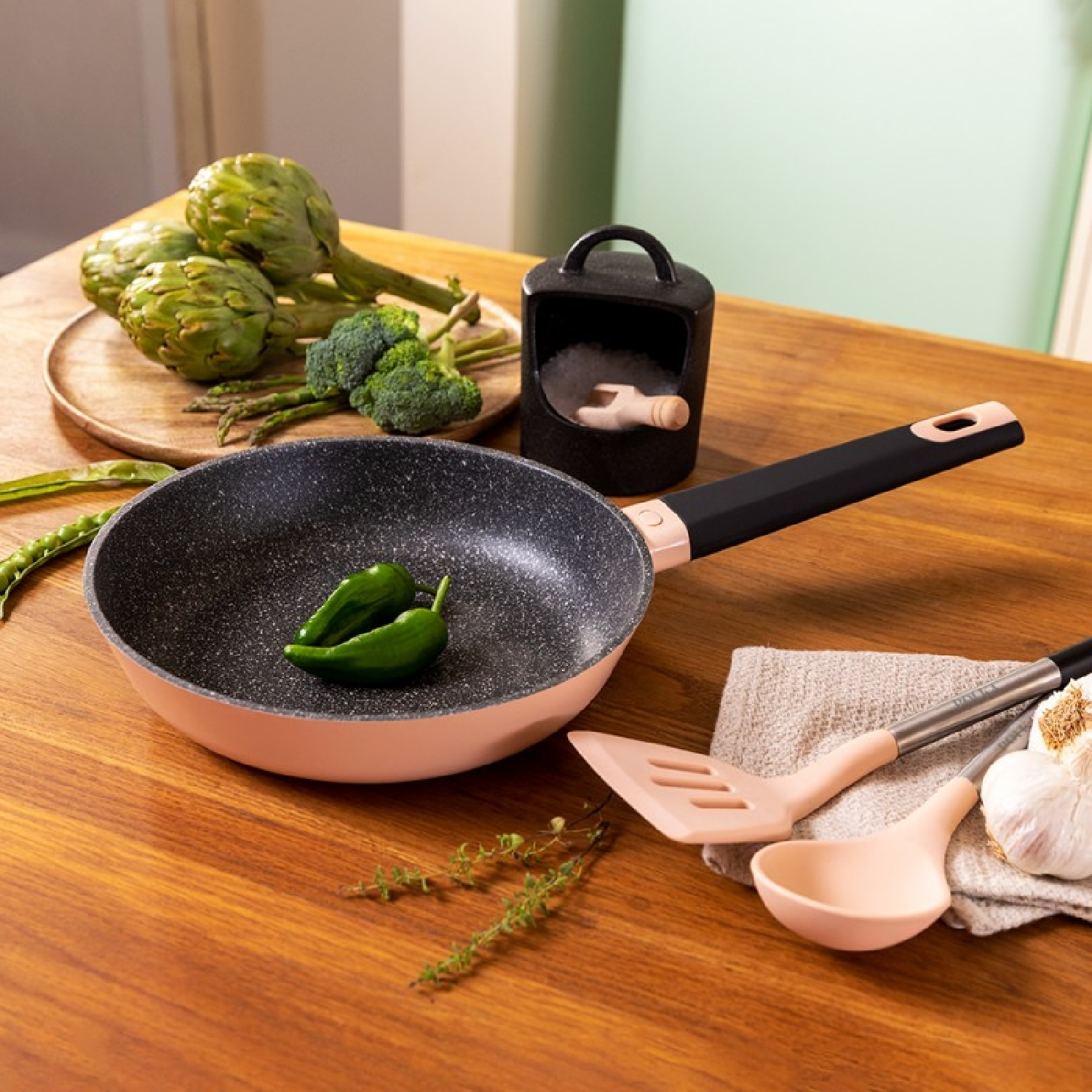 Een pan met een zwarte handgreep, wat groenten en keukengerei liggen op een houten tafel.