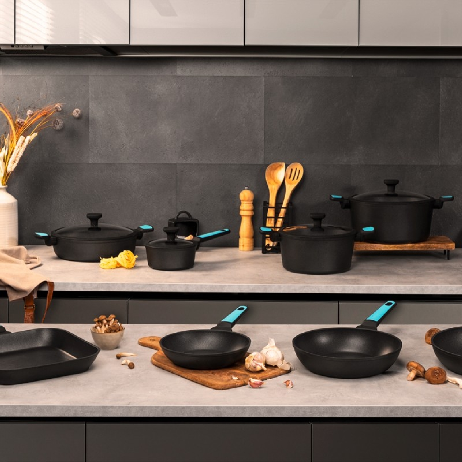 Cucina con varie pentole nere su un piano di lavoro grigio. Utensili in legno e funghi visibili.