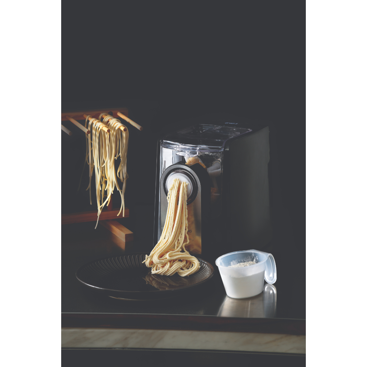 Macchina per la pasta che estrude spaghetti su un piatto. Pasta fresca appesa a un supporto di legno.
