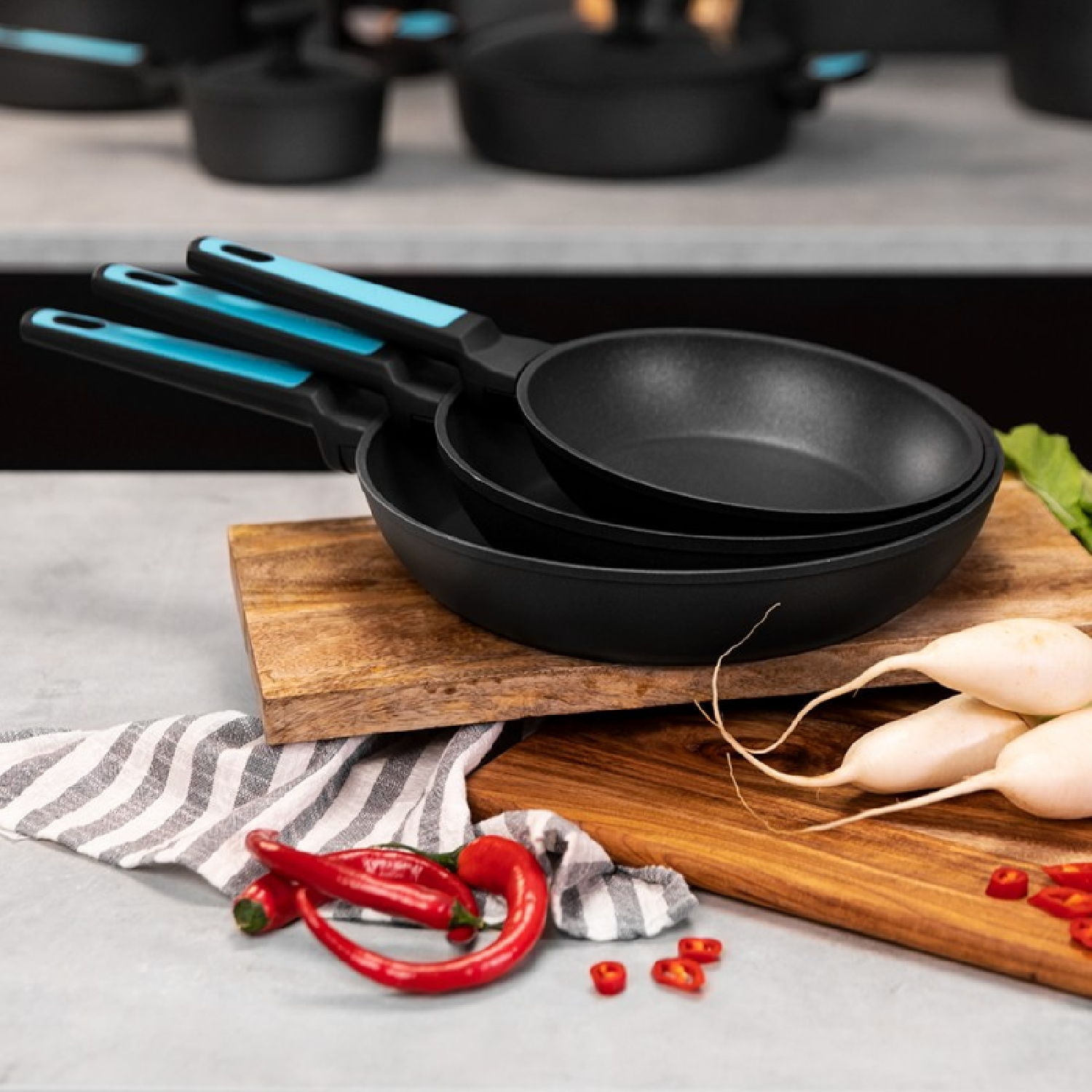 Set di tre padelle nere con manici blu, su un tagliere di legno con peperoni rossi.