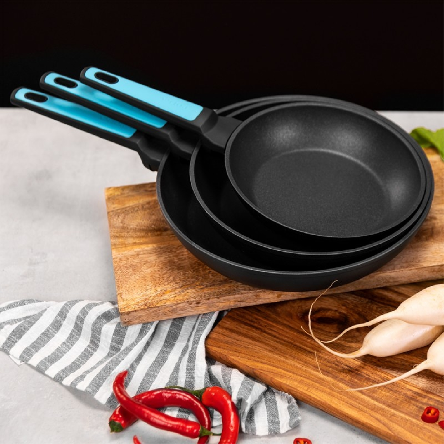 Set di tre padelle nere con manici blu su assi di legno con verdure.