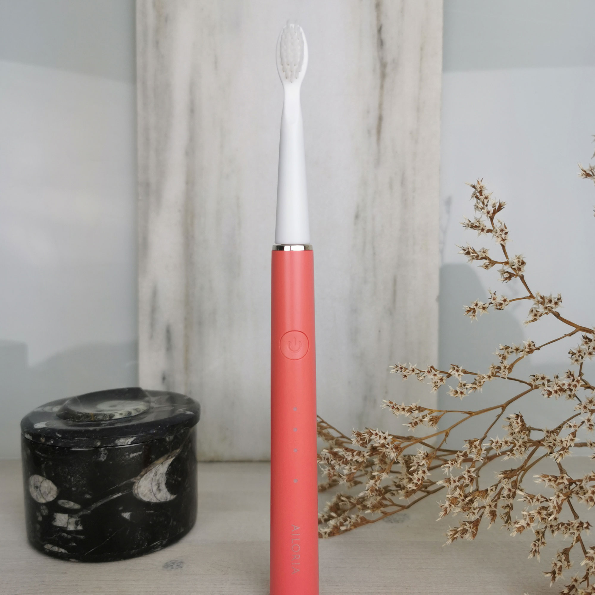 Cepillo de dientes eléctrico rosa con cerdas blancas sobre una superficie de madera, con piedra negra y flores.