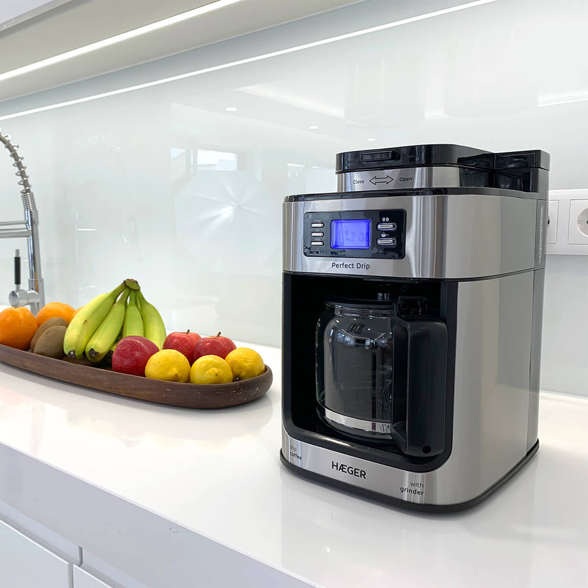 Srebrny ekspres do kawy z tacą owoców obok niego na białym blacie.