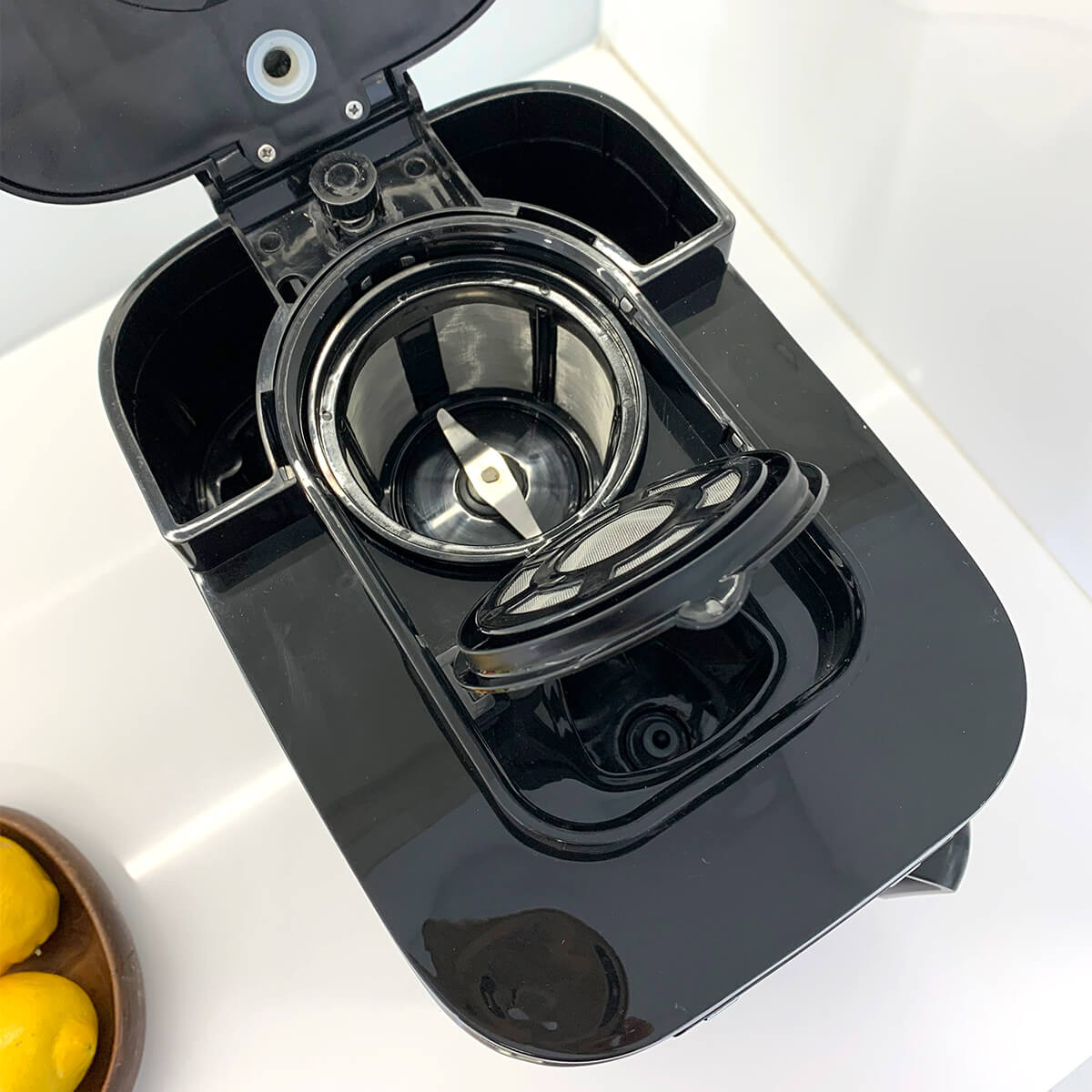 Czarny ekspres do kawy, widok z góry. Pokrywa otwarta, widoczny filtr i elementy wewnętrzne. Cytryny w tle.