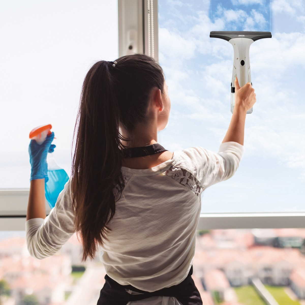 Mujer limpiando una ventana con un limpiacristales. Cielo azul de fondo. Lleva guantes y un spray.