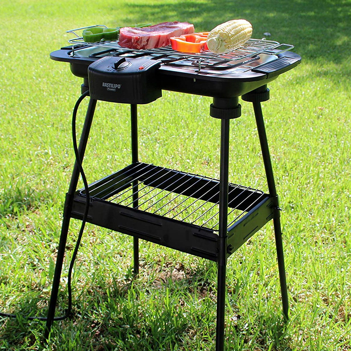 Parrilla eléctrica con carne y verduras. Está sobre un soporte en un césped.