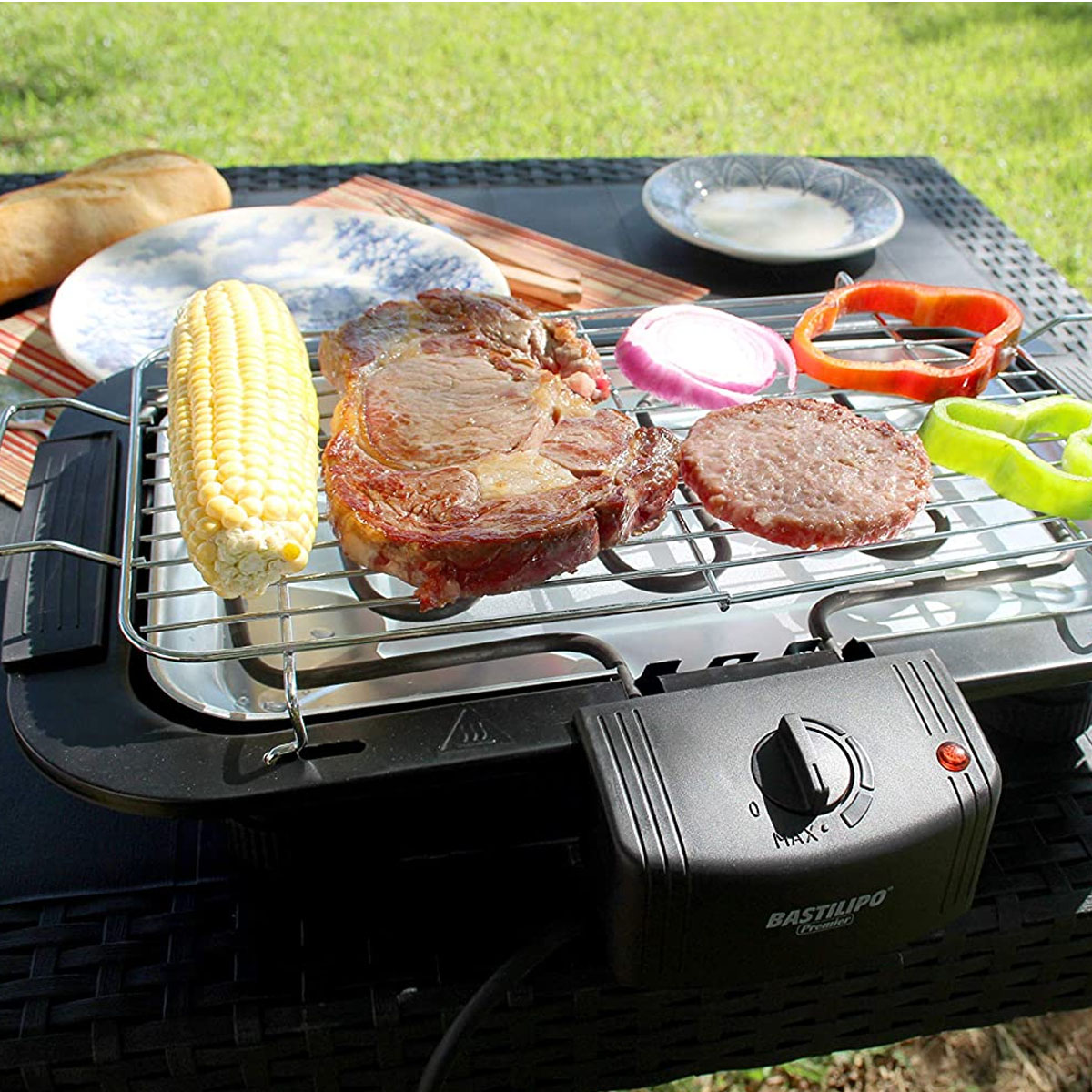 Una parrilla eléctrica con comida encima, incluyendo filete, maíz y verduras. Al aire libre.