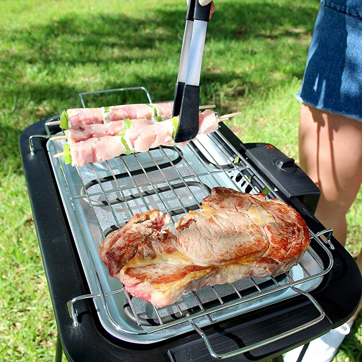 Una persona asando carne en una pequeña parrilla eléctrica en un entorno al aire libre con césped.