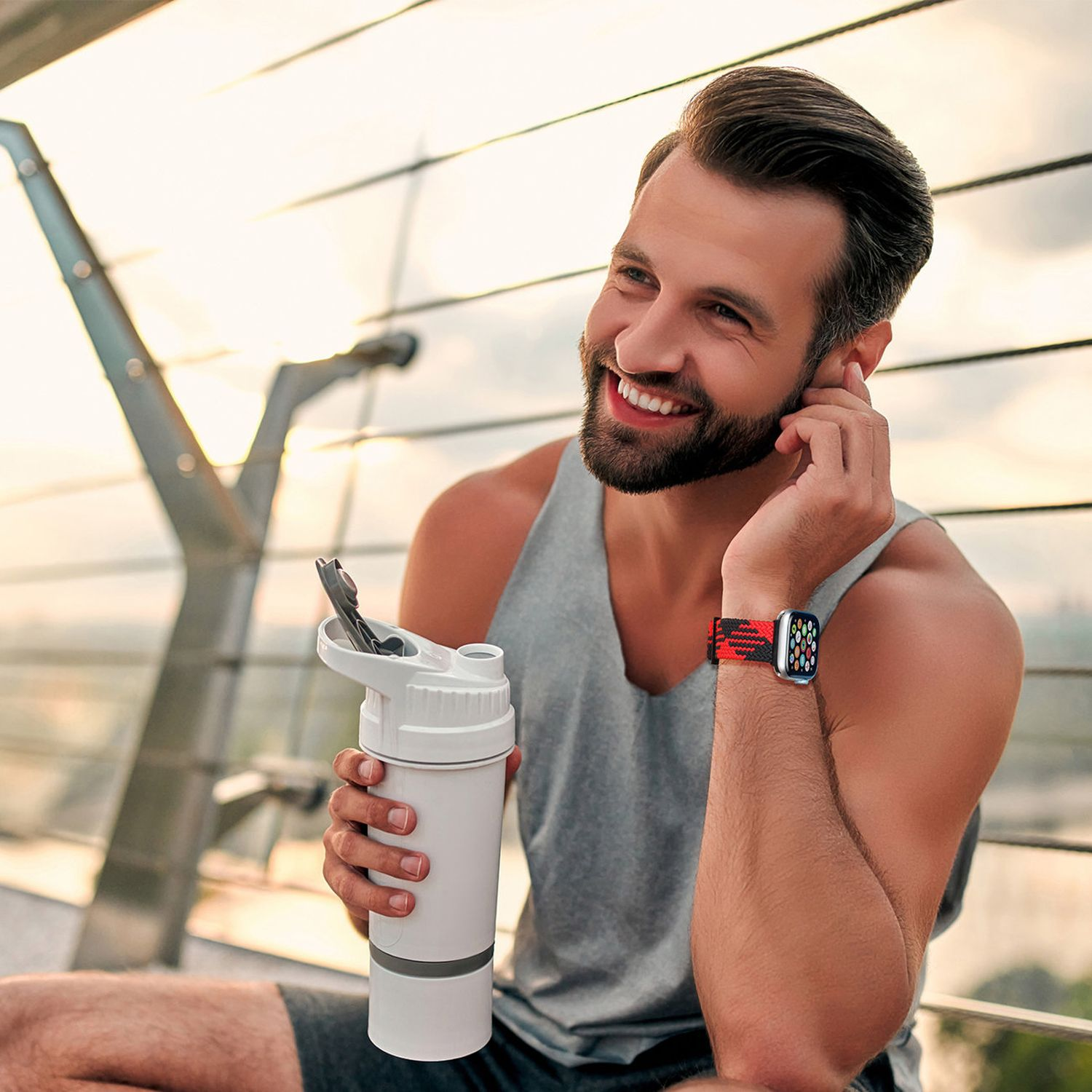 Mann mit Smartwatch und Tanktop, lächelnd. Hält eine Wasserflasche. Draußen mit verschwommenem Hintergrund.