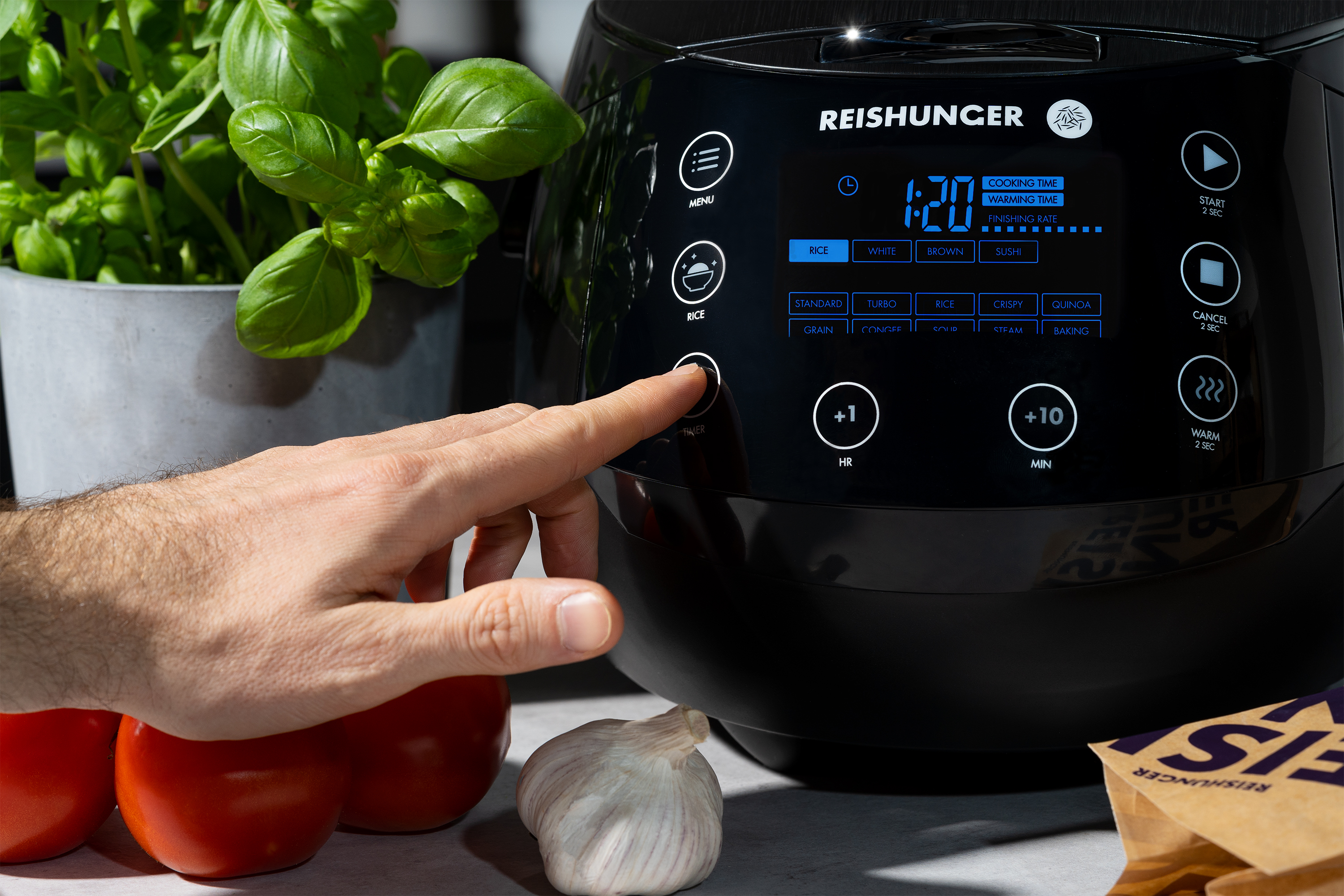 Eine Hand drückt einen Knopf auf einem schwarzen Reiskocher neben Tomaten und Basilikum.
