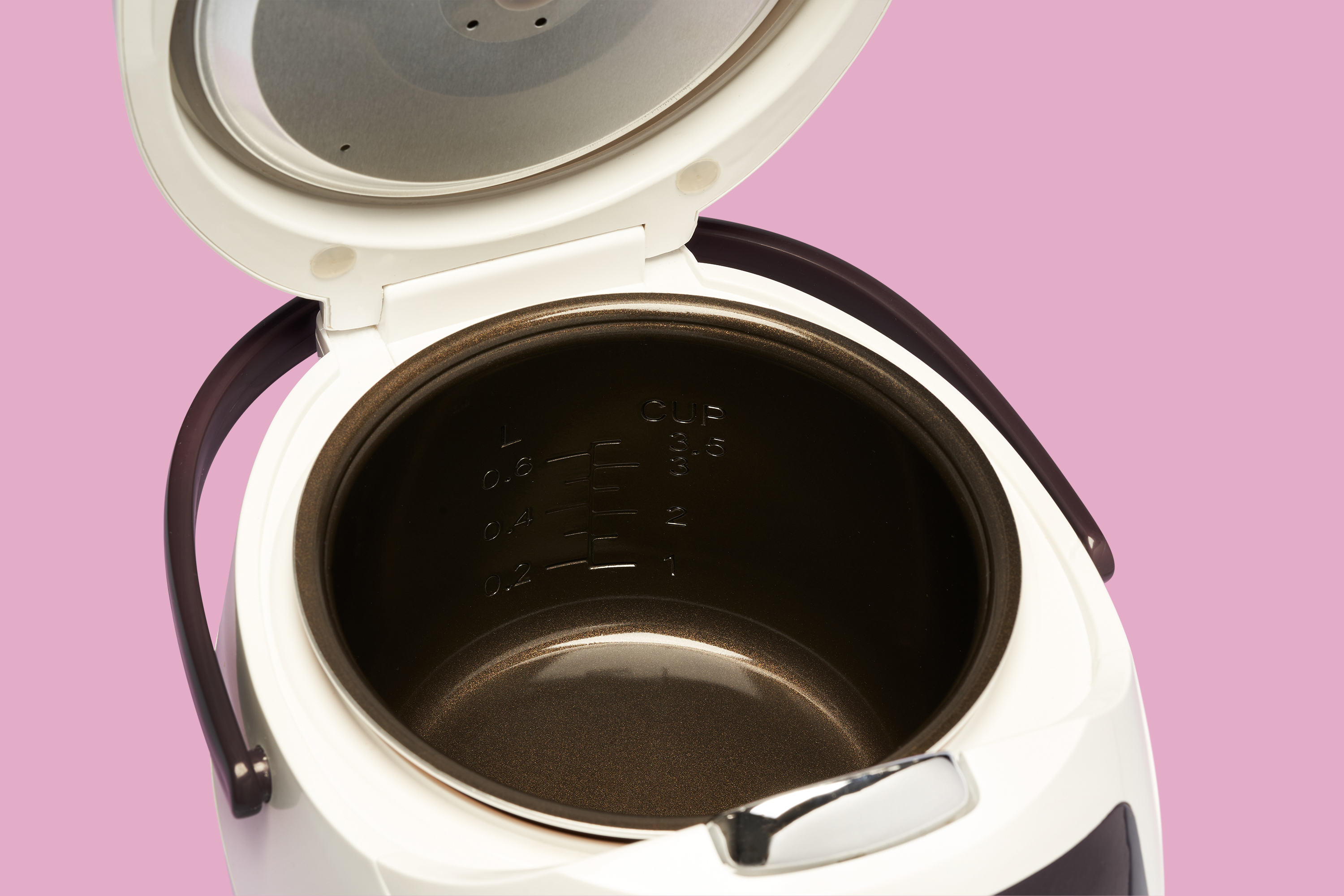 Inneres eines weißen Reiskochers mit offenem Deckel. Der Innenraum ist braun. Rosa Hintergrund.