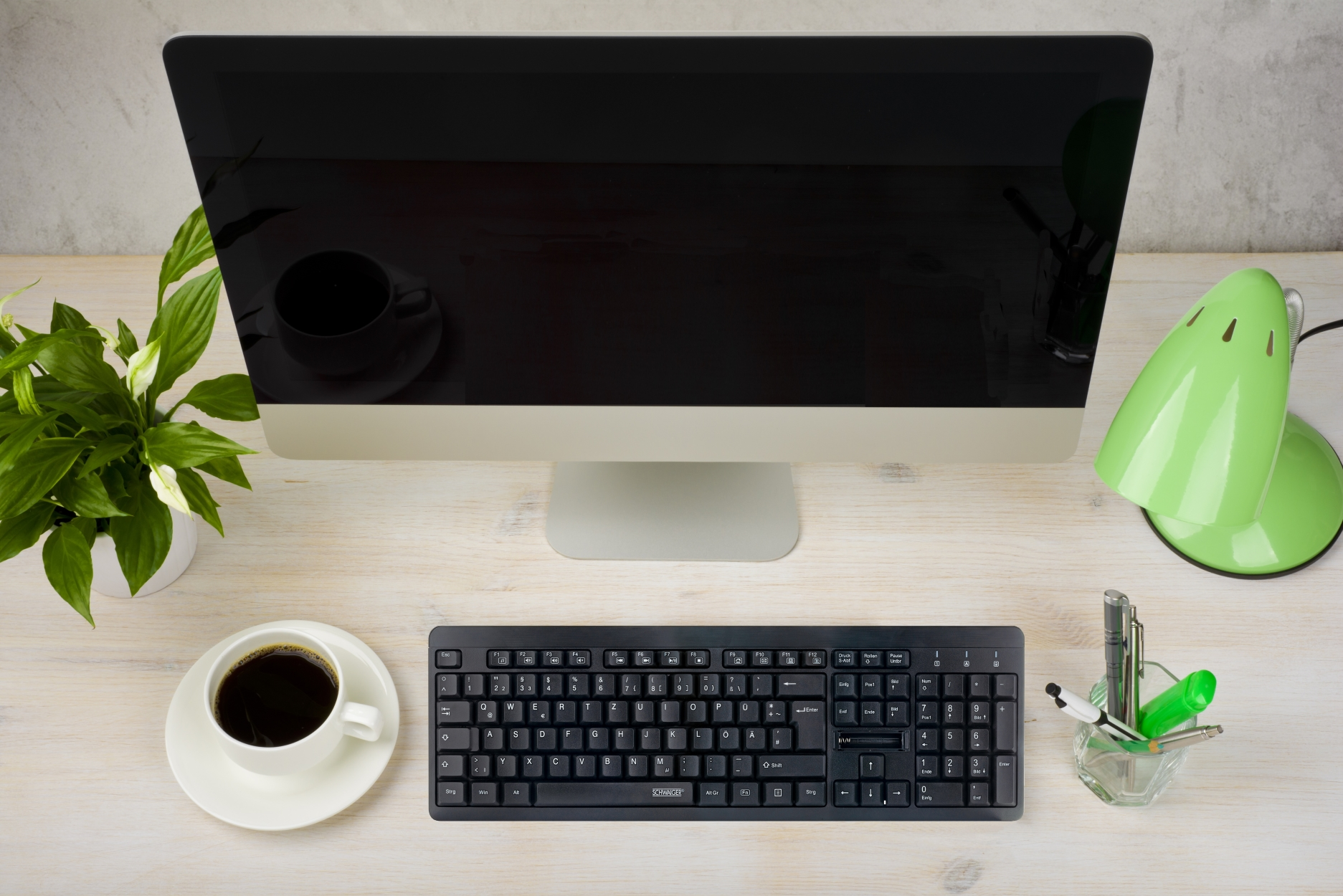 Una scrivania con monitor, tastiera, caffè, pianta e una lampada da scrivania verde.