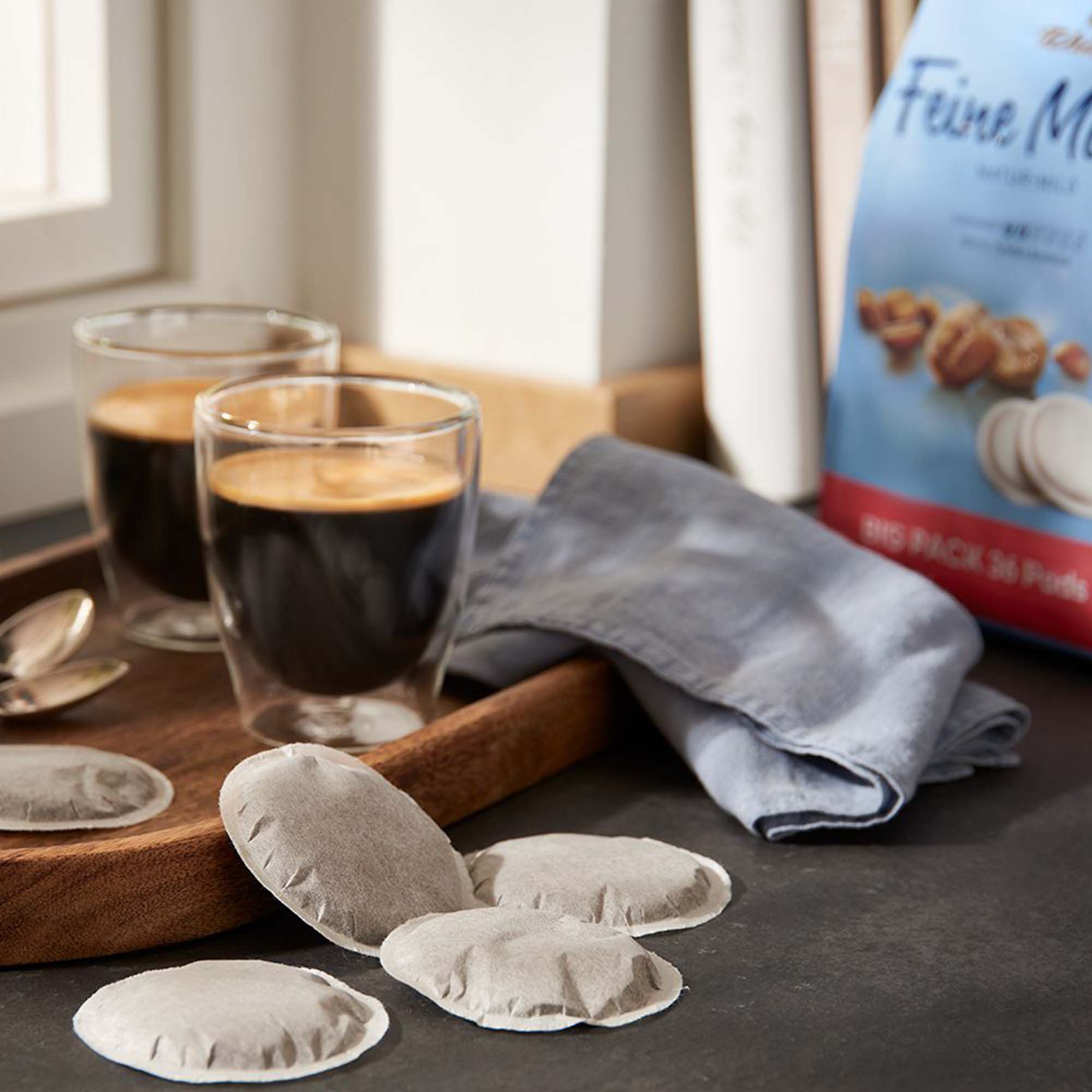 Zwei Kaffeegläser und Kaffeepads auf einem Holztablett. Bücher sind im Hintergrund.