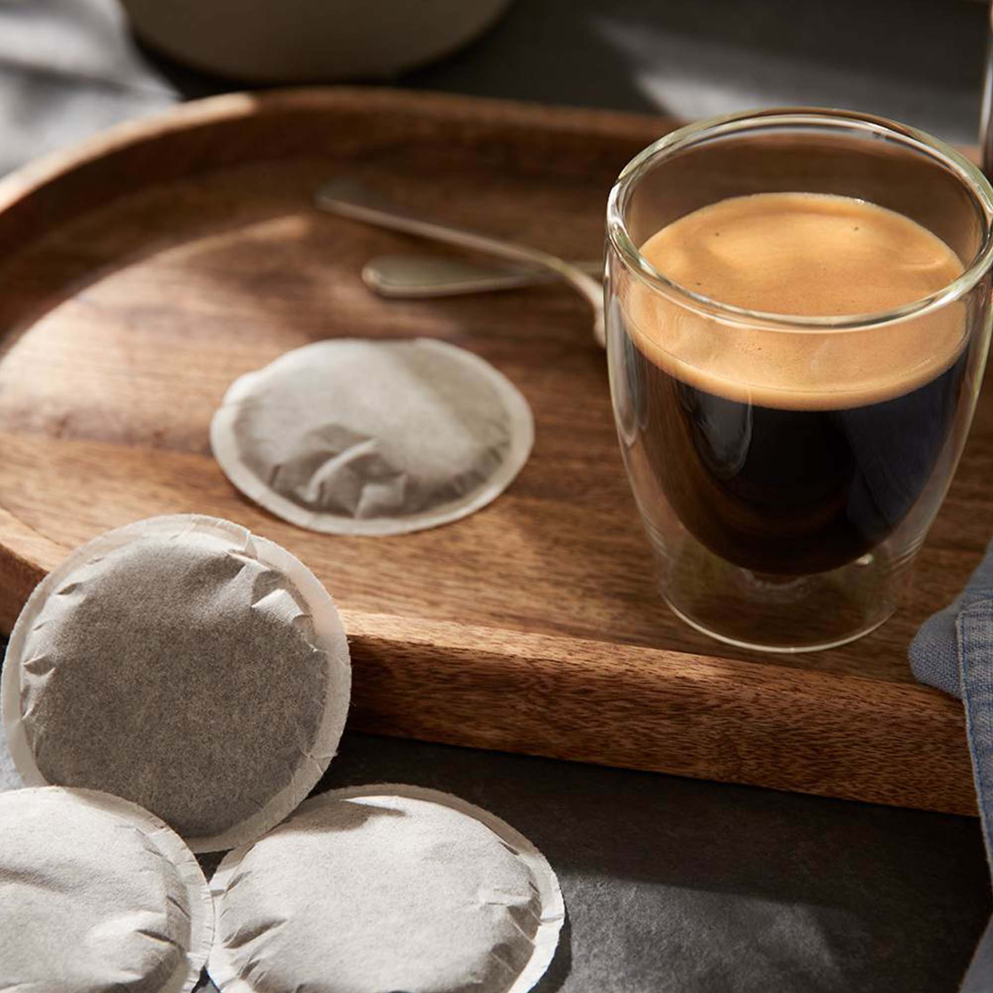 Kaffee in einem klaren Glas, mit Kaffeepads auf einem Holztablett auf weißem Grund.