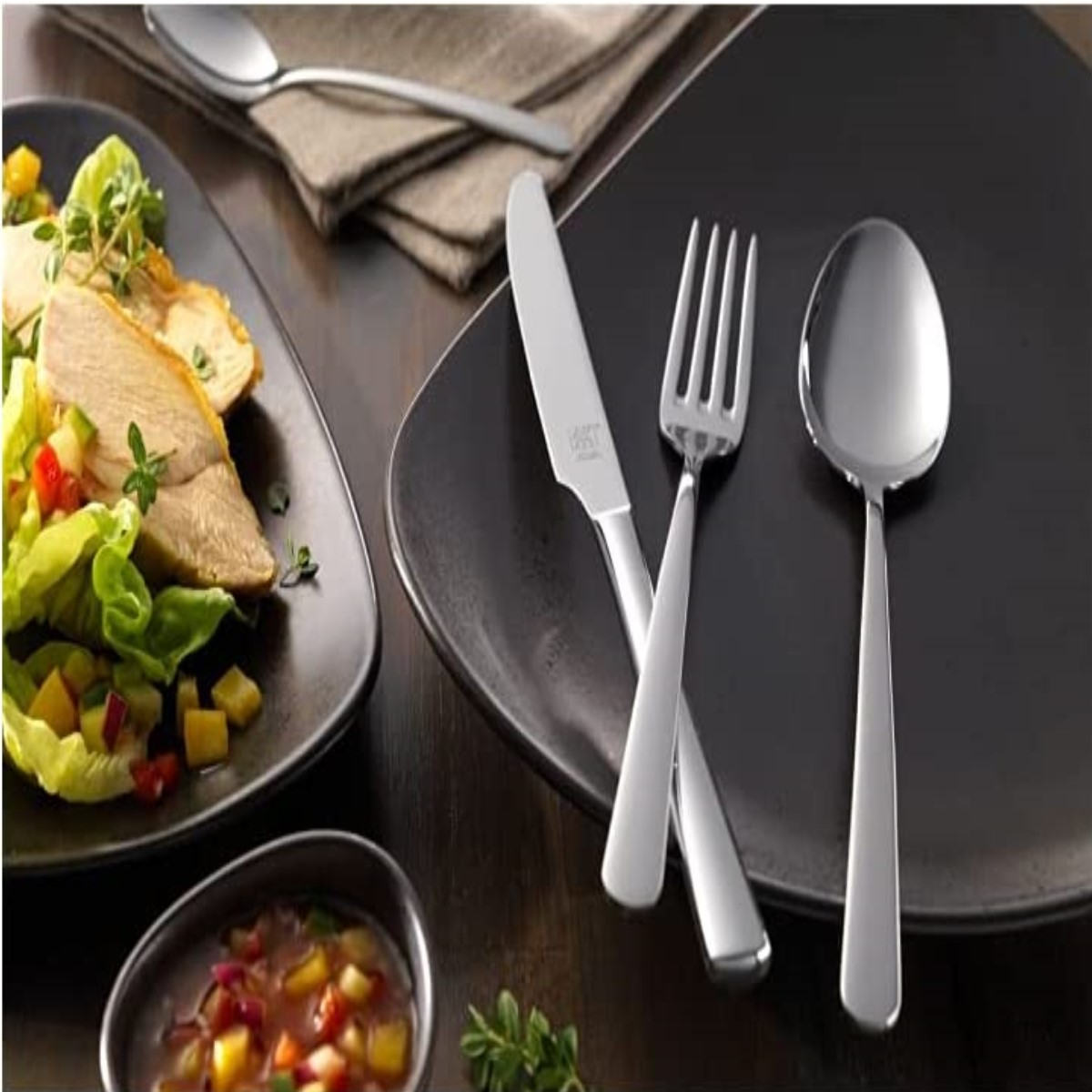Teller mit Essen und Besteck auf einem dunklen Holztisch. Salat mit Dressing, Gabel, Messer und Löffel.