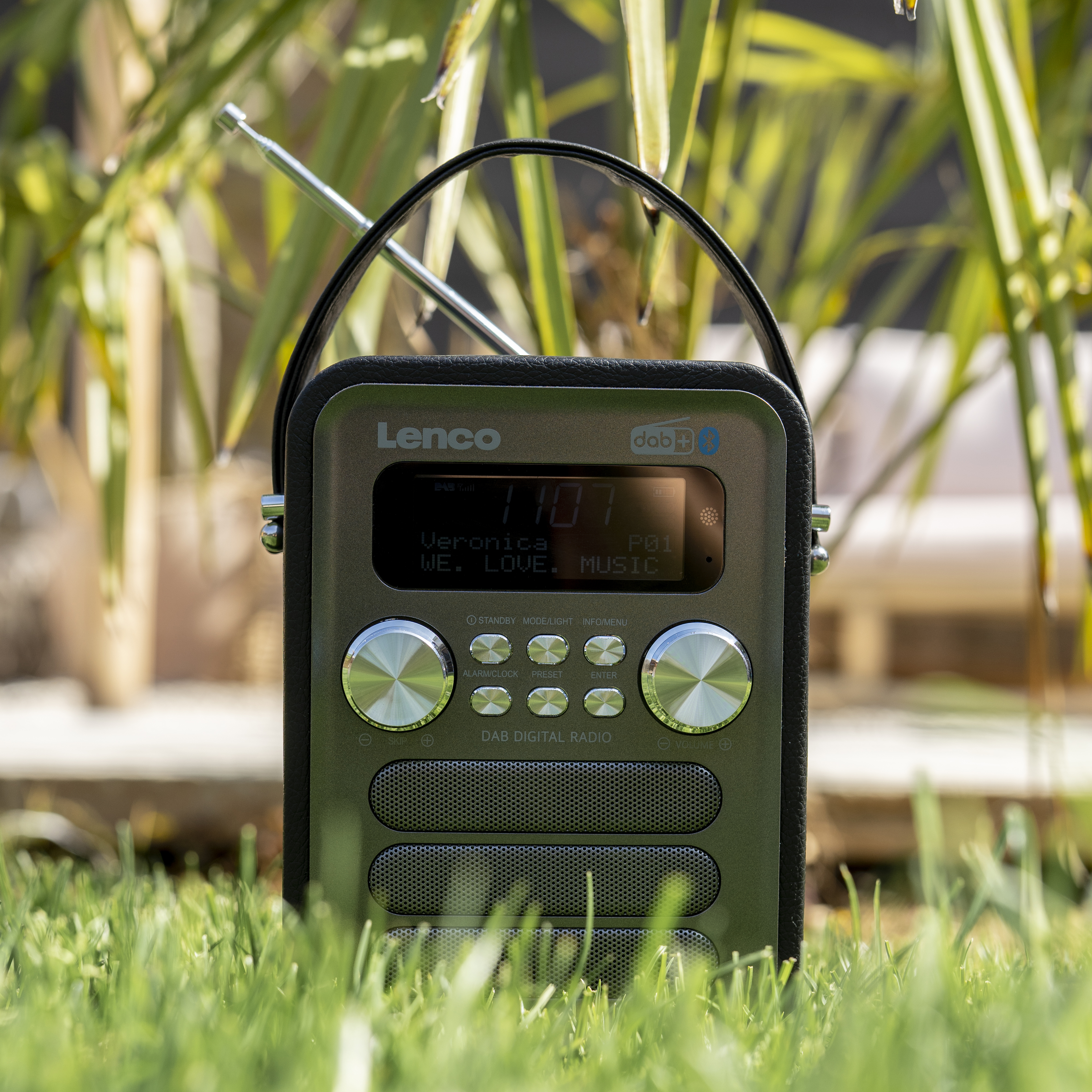 Przenośne radio cyfrowe DAB na zielonej trawie. Posiada ekran, przyciski i antenę.