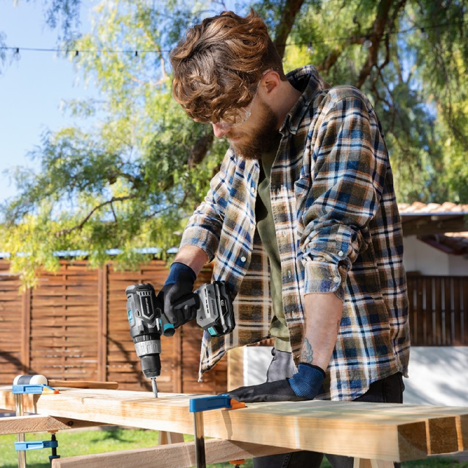 Mężczyzna wierci drewno na zewnątrz. Ma na sobie koszulę w kratę i rękawiczki. Drewniany stół jest na pierwszym planie.