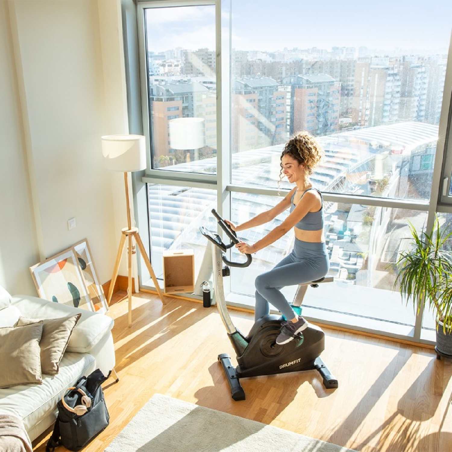 Donna su cyclette in una stanza luminosa con vista sulla città. Un divano bianco e una lampada da terra sono visibili.