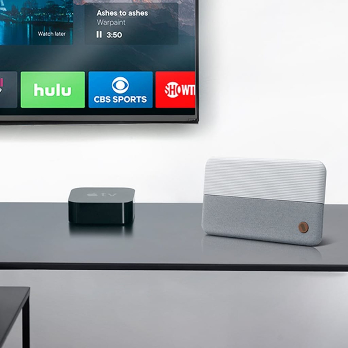 A TV displaying streaming service logos, with a black Apple TV and a gray and white speaker on the table.