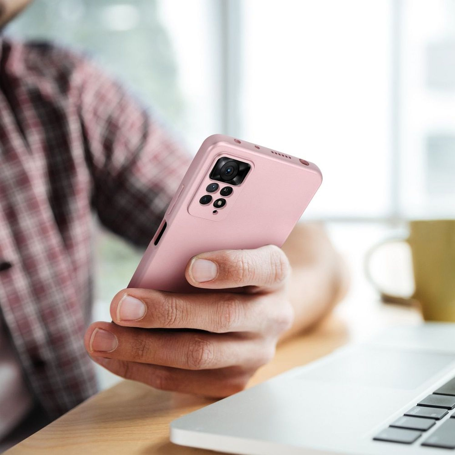 Person hält pinkes Telefon mit Kamera auf Schreibtisch mit Laptop und Becher.