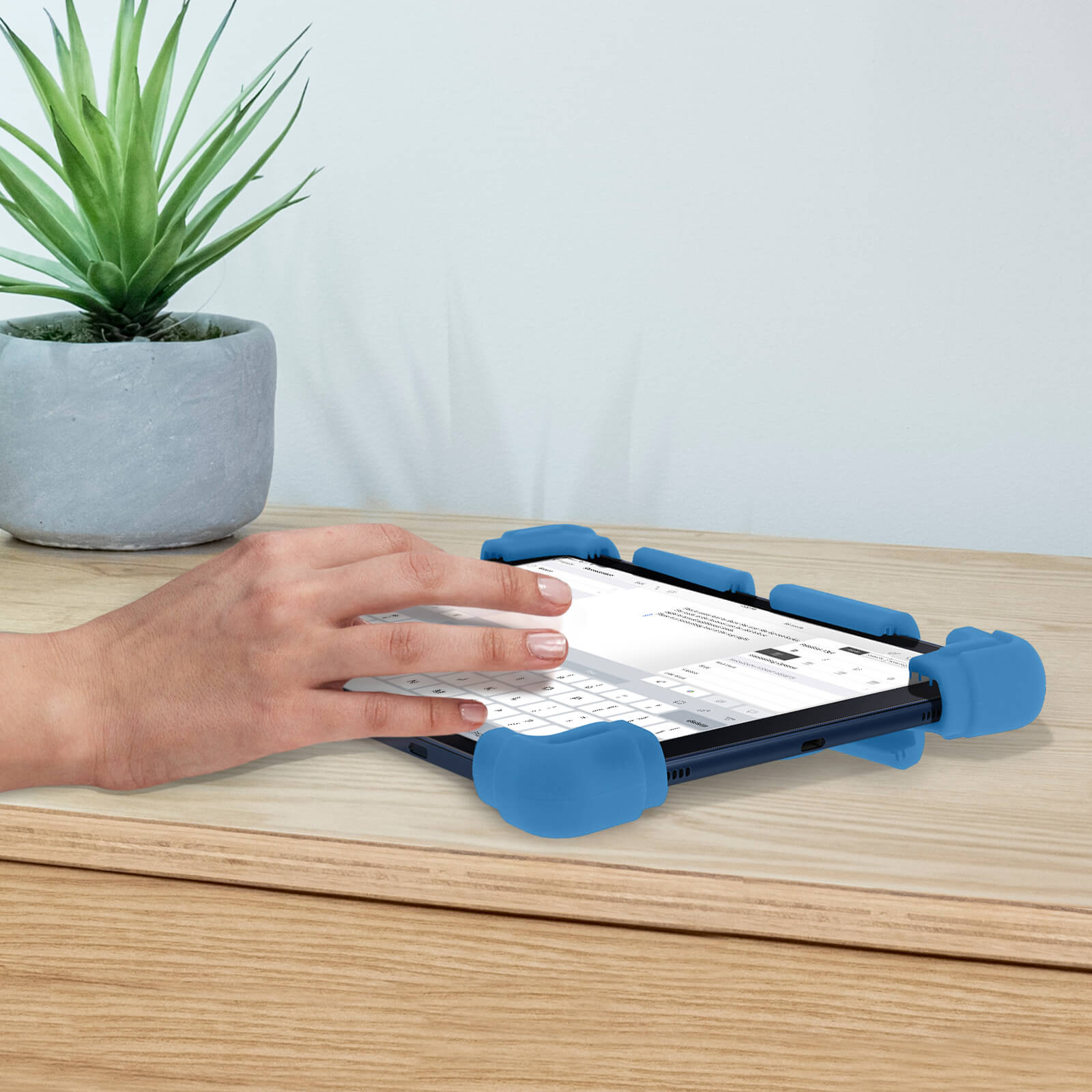 Eine Hand tippt auf einem Tablet mit blauem Schutzgehäuse. Eine Pflanze steht auf einem Holztisch.