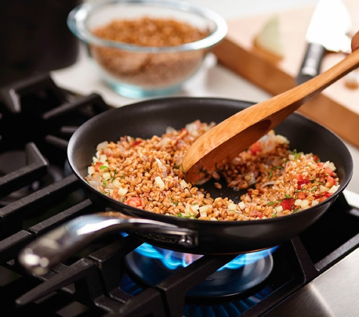 Sartén en una estufa de gas con comida cocinándose, removida con una cuchara de madera. Un cuenco en el fondo.
