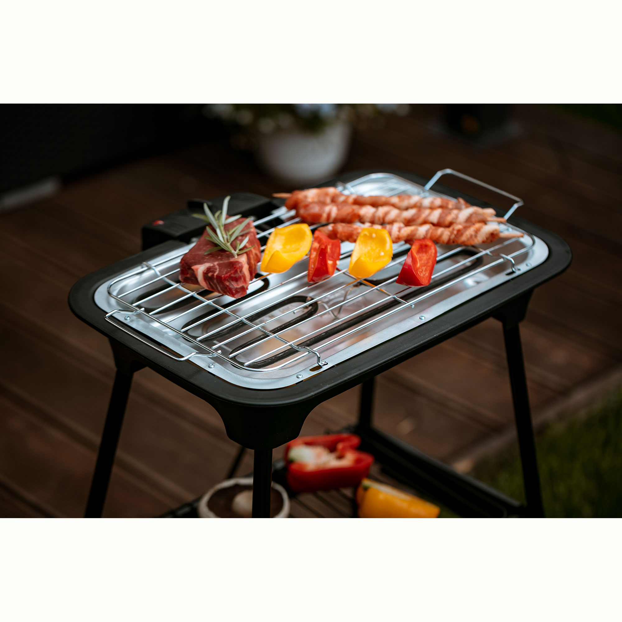 Grill mit Fleisch und Gemüse auf Holzterrasse, grüner Hintergrund.