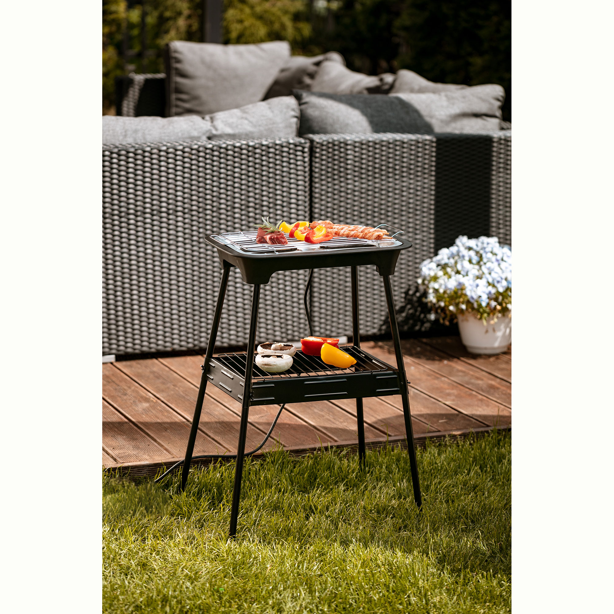 Schwarzer Grill mit Essen darauf, auf einer Holzterrasse mit grünem Gras und einem grauen Sofa im Hintergrund.