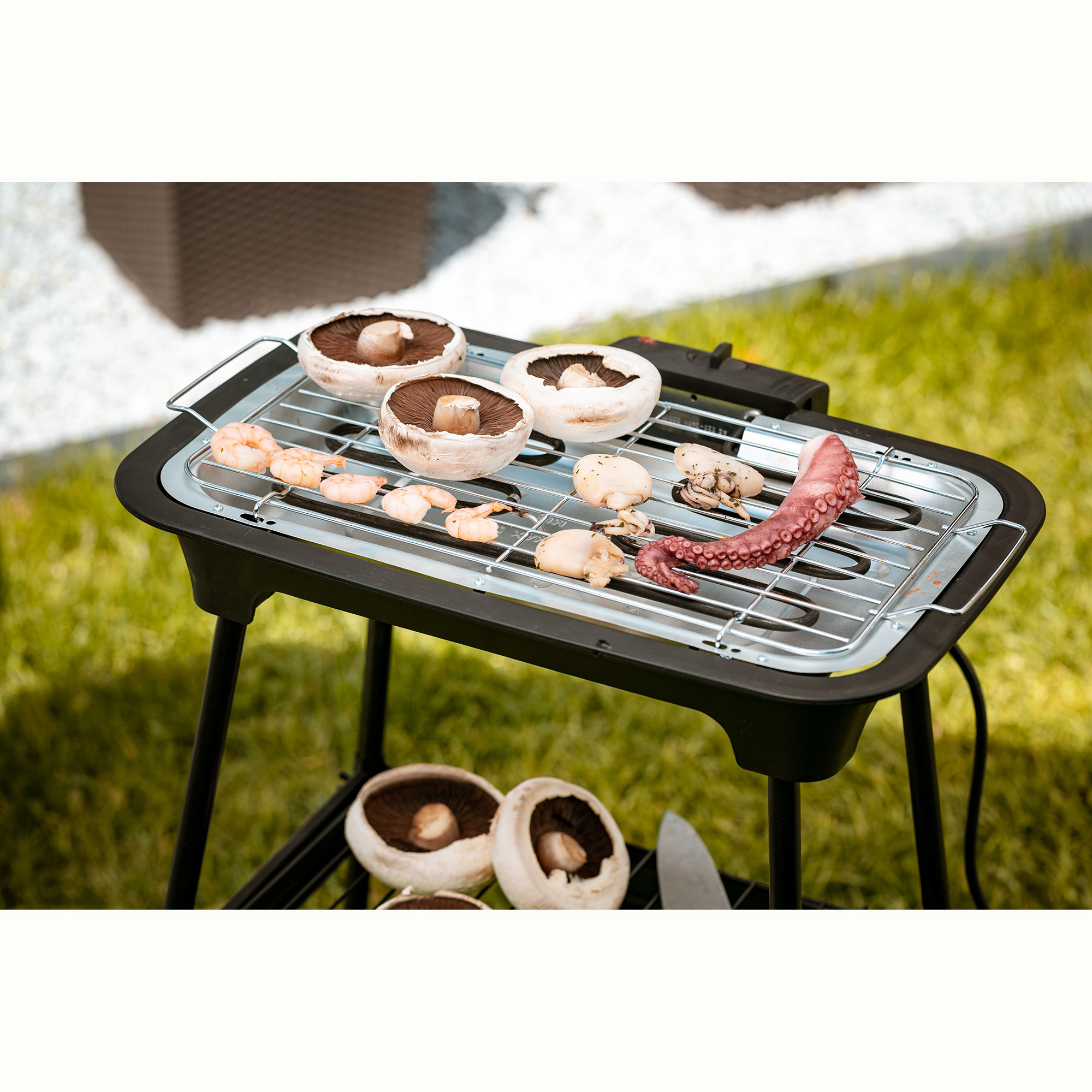 Elektrischer Außengrill mit Essen darauf, Pilze, Garnelen und Oktopus. Grasiger Hintergrund.
