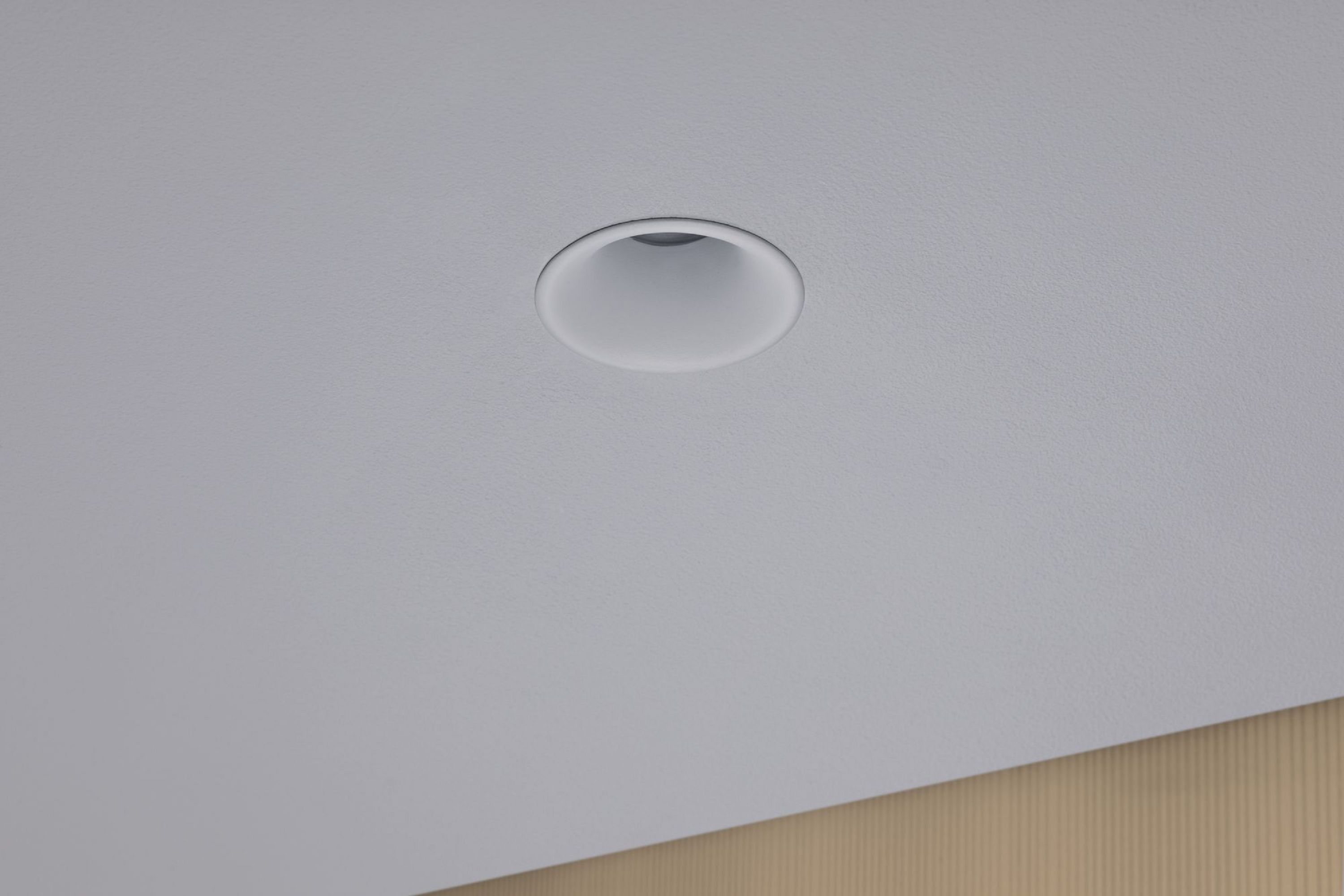 Ein Einbaustrahler, kreisförmig, in einer weißen Decke, mit einer beigen Wand darunter.