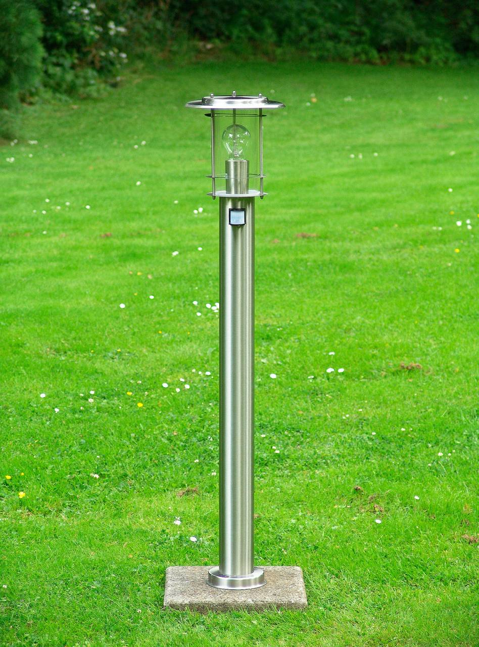 Eine moderne Edelstahl-Gartenleuchte steht auf einem Betonfundament in einem grasbewachsenen Garten.