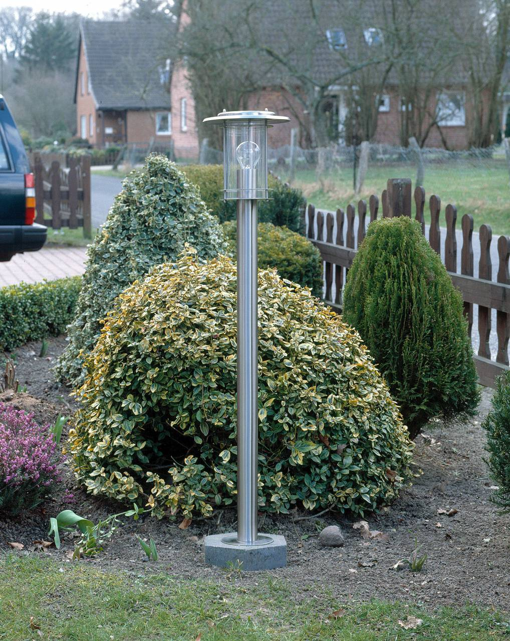 Ein moderner Außenleuchter steht im Garten vor Büschen und Bäumen.