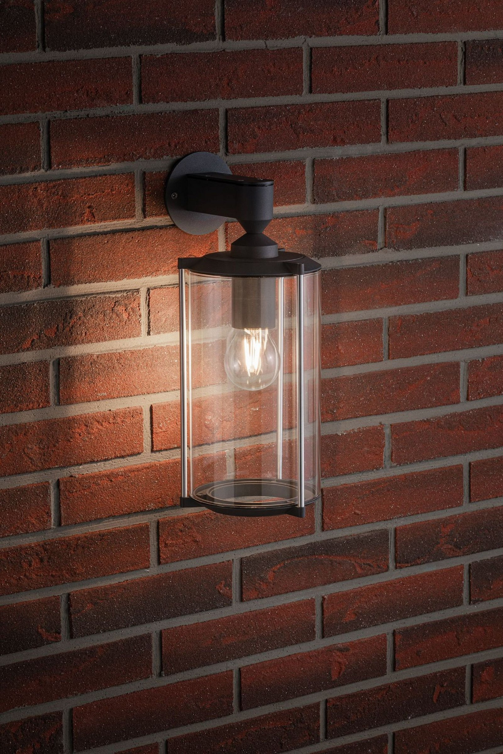Eine schwarze Außenlampe an einer roten Backsteinmauer. Die Lampe hat einen klaren Glaszylinder und eine Glühbirne.