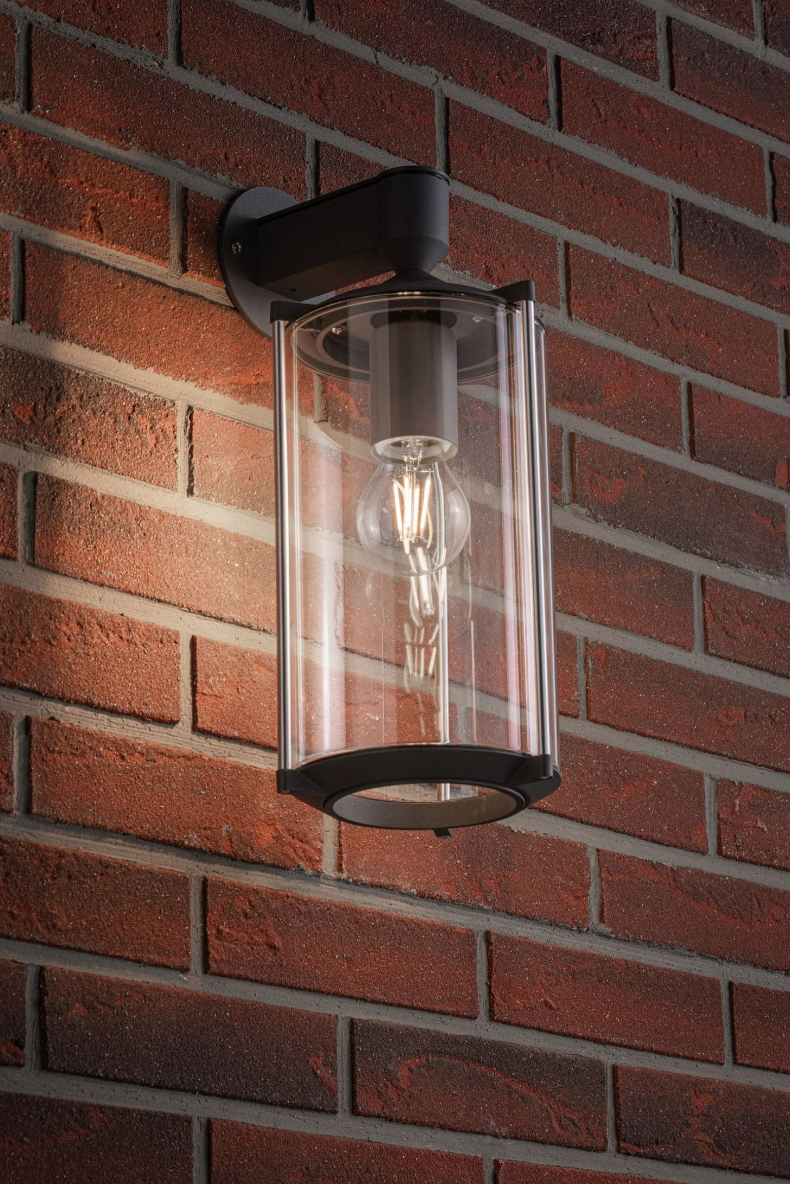 Außenwandleuchte an einer roten Backsteinmauer. Die Leuchte hat ein klares Glasgehäuse.