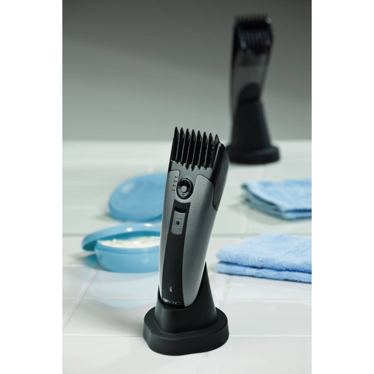 Szara golarka elektryczna na stojaku ładującym. Druga golarka i kosmetyki w tle.