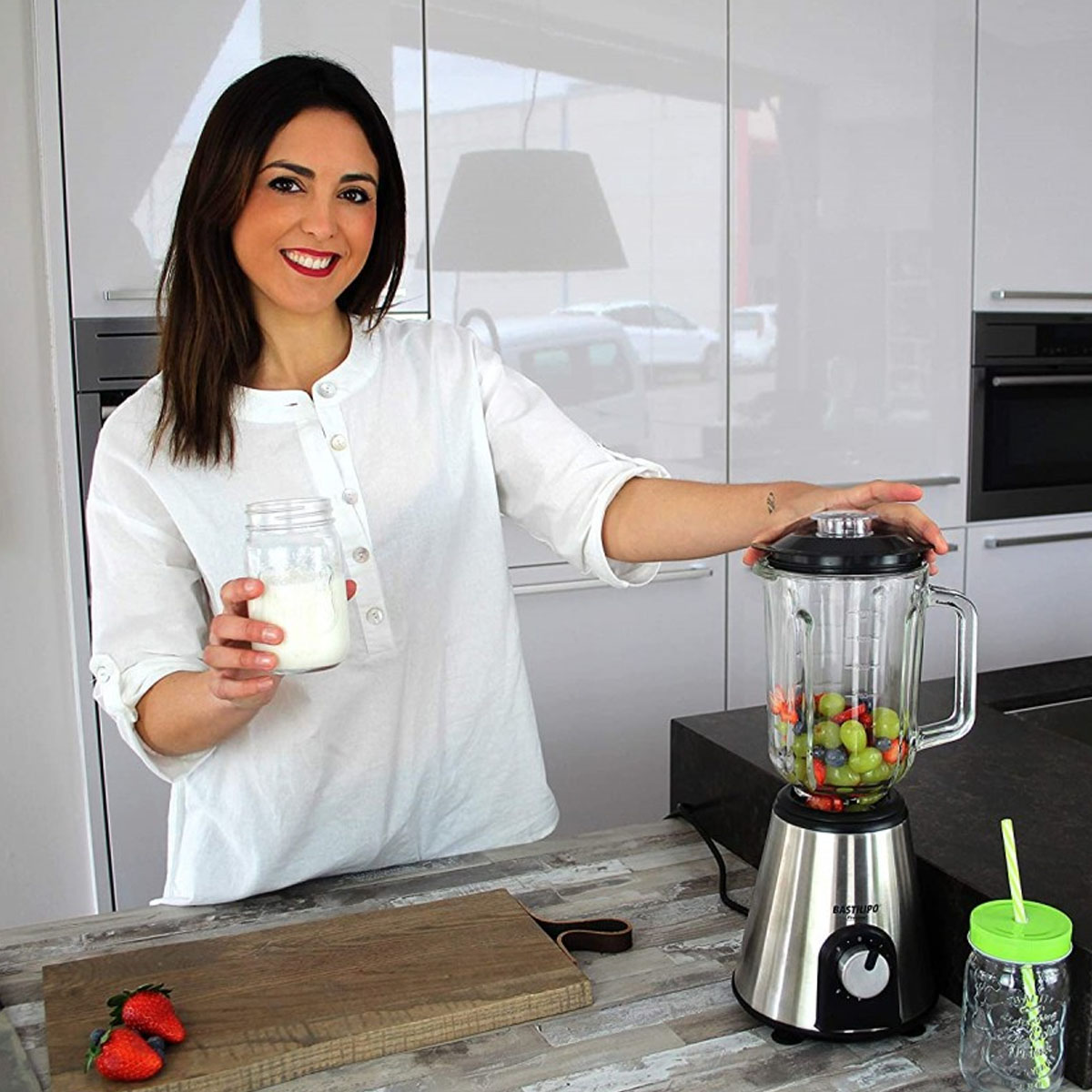 Una mujer sostiene un frasco de leche y una licuadora con fruta. Una escena de cocina.