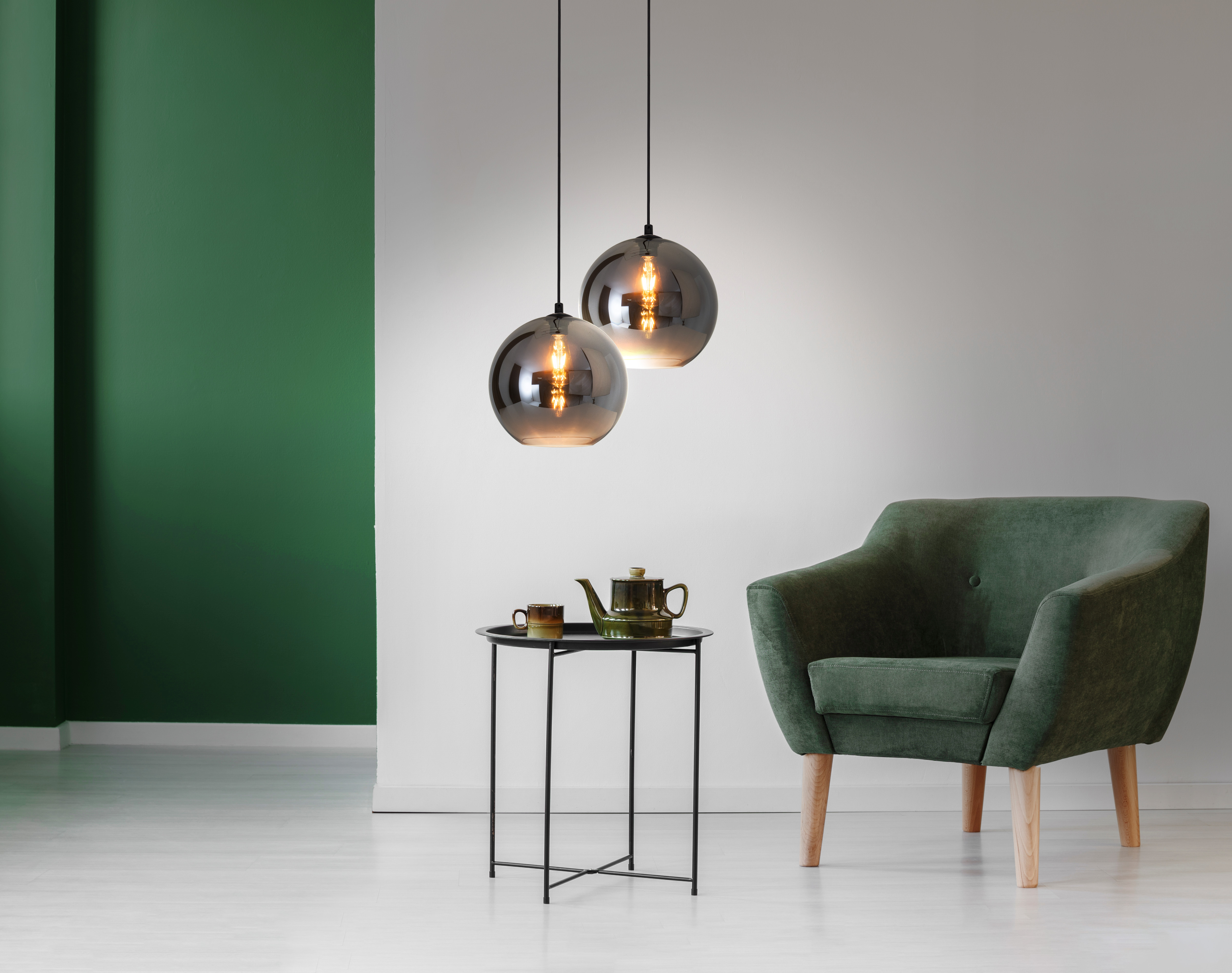 Deux lampes sphériques en verre suspendues au-dessus d'une petite table noire avec un fauteuil vert et un mur vert.